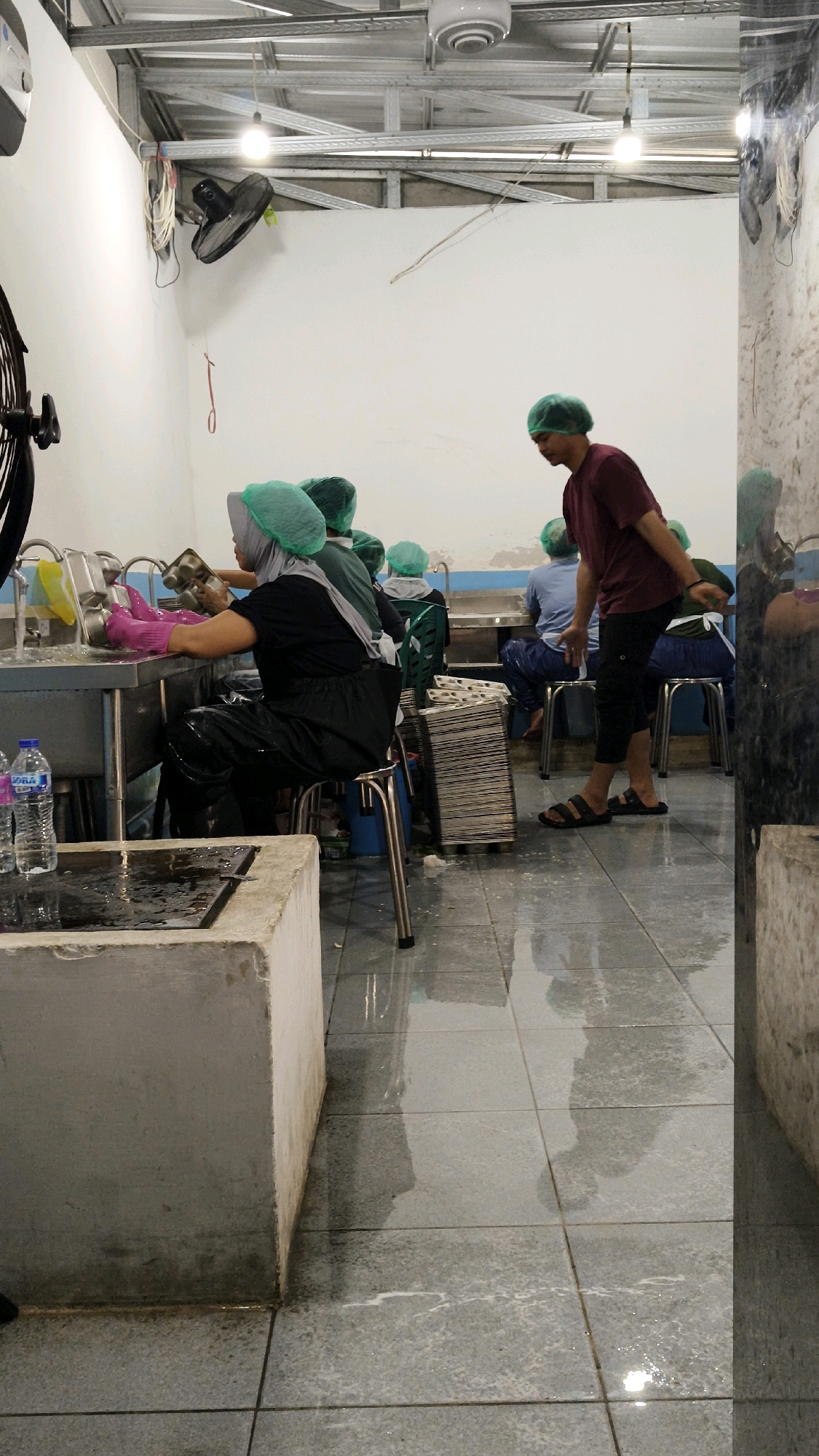 The image depicts an indoor scene, possibly a workshop or food processing area, in Sambipondok, Indonesia. The environment is utilitarian, with a metal ceiling structure and white walls. The floor is tiled and appears wet, reflecting the overhead lighting.
In the foreground, on the left, is a concrete structure with a damp surface. To its right, a stainless-steel workstation with a sink is visible. A woman wearing a green hairnet and pink gloves is seated at this workstation, appearing to wash or clean metal trays. Two plastic bottles of water are on the workstation.
Several other individuals, all wearing green hairnets, are present. One person, wearing a maroon t-shirt and dark pants, stands facing away from the camera, seemingly interacting with someone out of frame or observing the scene. Other individuals are seated or standing in the background, engaged in their tasks, though their actions are less clear. A stack of metal trays or containers is positioned to the right of the seated woman. An industrial fan is mounted on the wall to the left. The overall atmosphere suggests a working environment where hygiene is likely a priority, given the hairnets and the cleaning activities.
