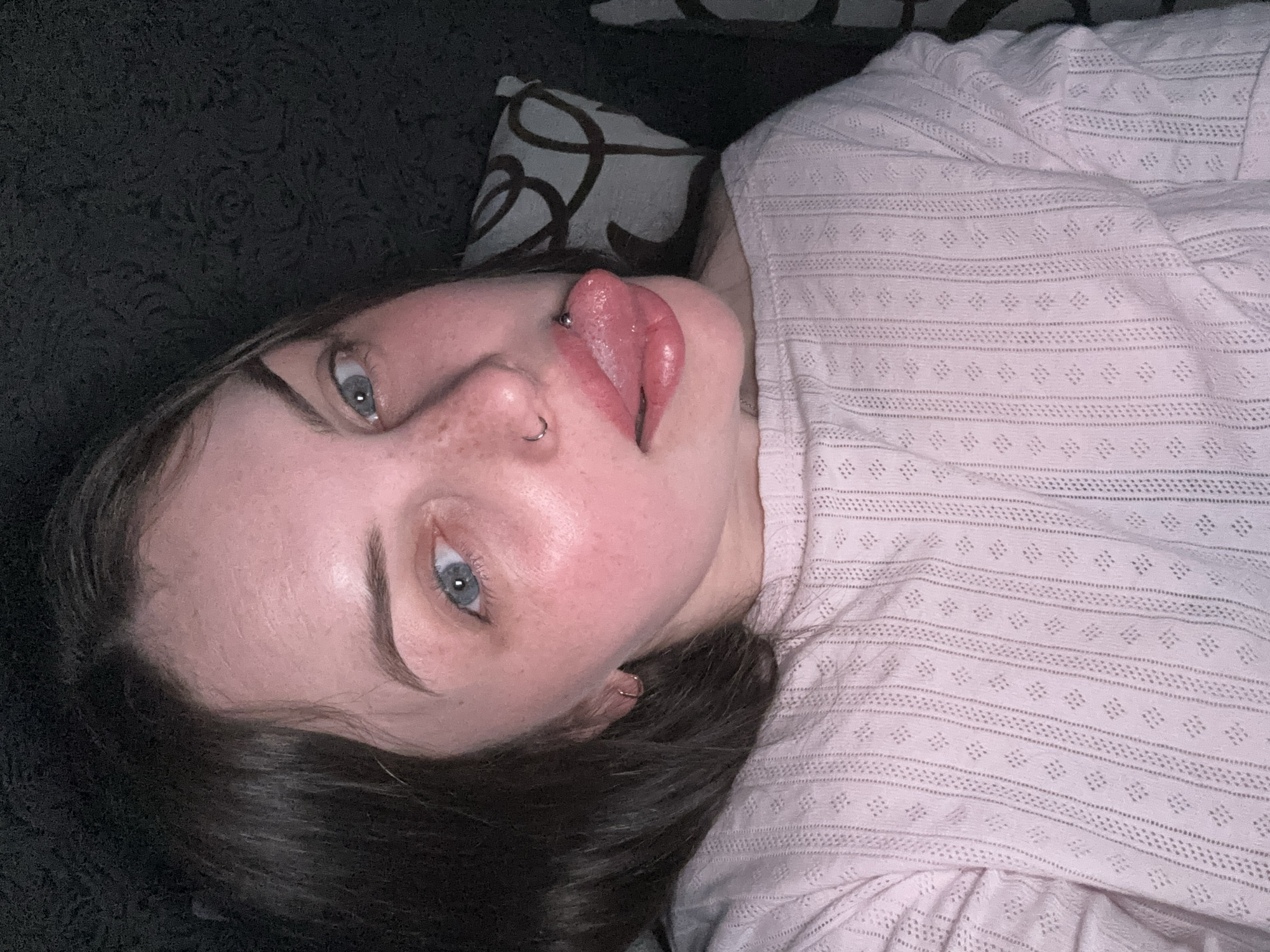 A young woman with pale skin, blue eyes, and dark hair is lying down. She has a nose piercing and a split tongue piercing. She is wearing a pink patterned shirt. She is lying on a dark-colored, patterned surface next to a patterned pillow. The location is not discernible from the image, and there is no indication it is in Chornomorskoe, Ukraine.