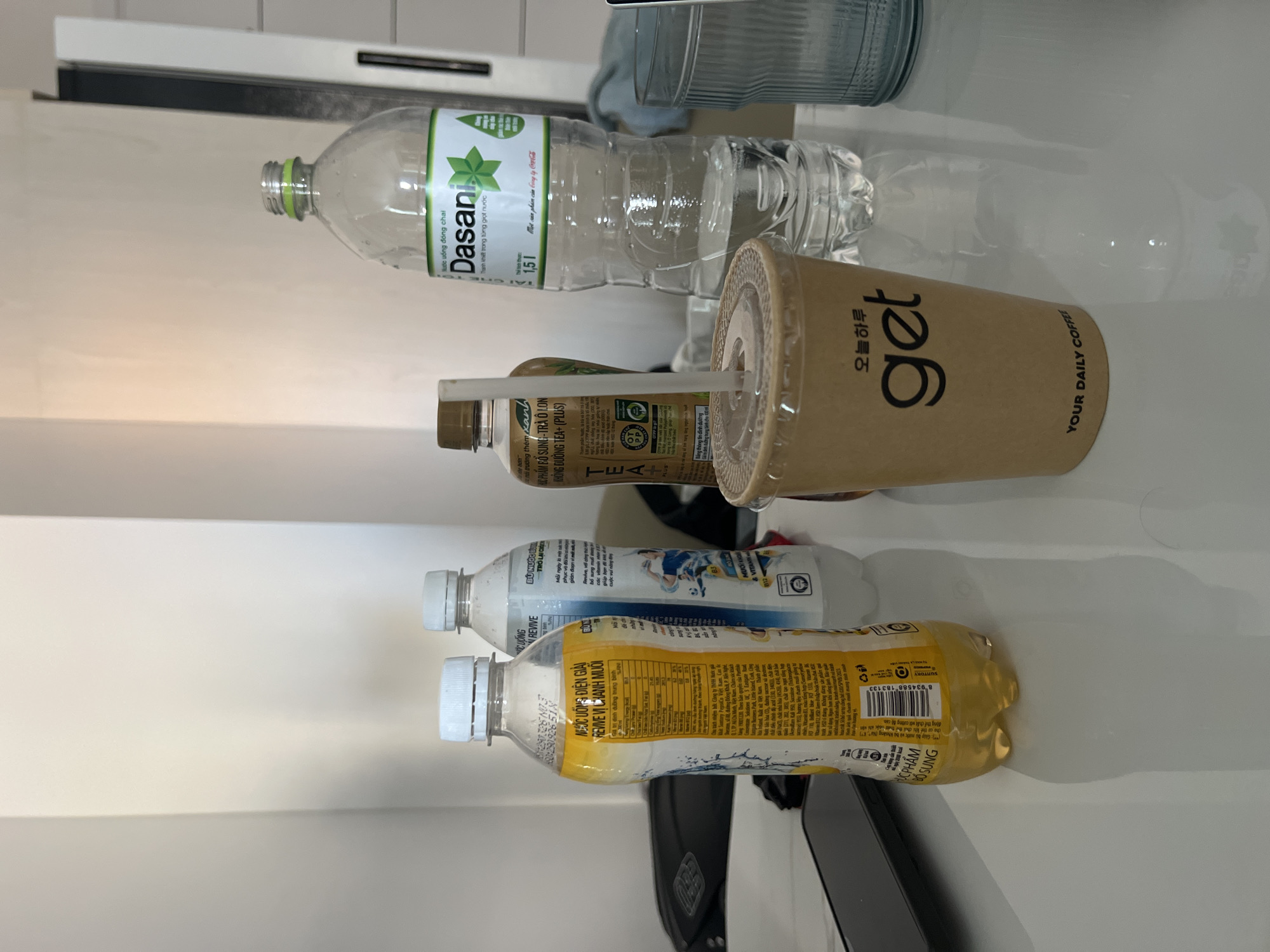 The image presents a flat lay arrangement of various beverage containers on a white surface. The objects include a bottle of Dasani water, a "get YOUR DAILY COFFEE" beverage, a bottle labeled "TEA+ (PLUS)", a Revive bottle with a sports player on the label, and another Revive bottle. The text in Vietnamese on the labels suggests the scene is in Vietnam, potentially in the city of Bắc Nha Trang. The containers are of varying sizes and states of fullness. The setting is indoors, presumably a refrigerator or storage area due to the clean, white surface.