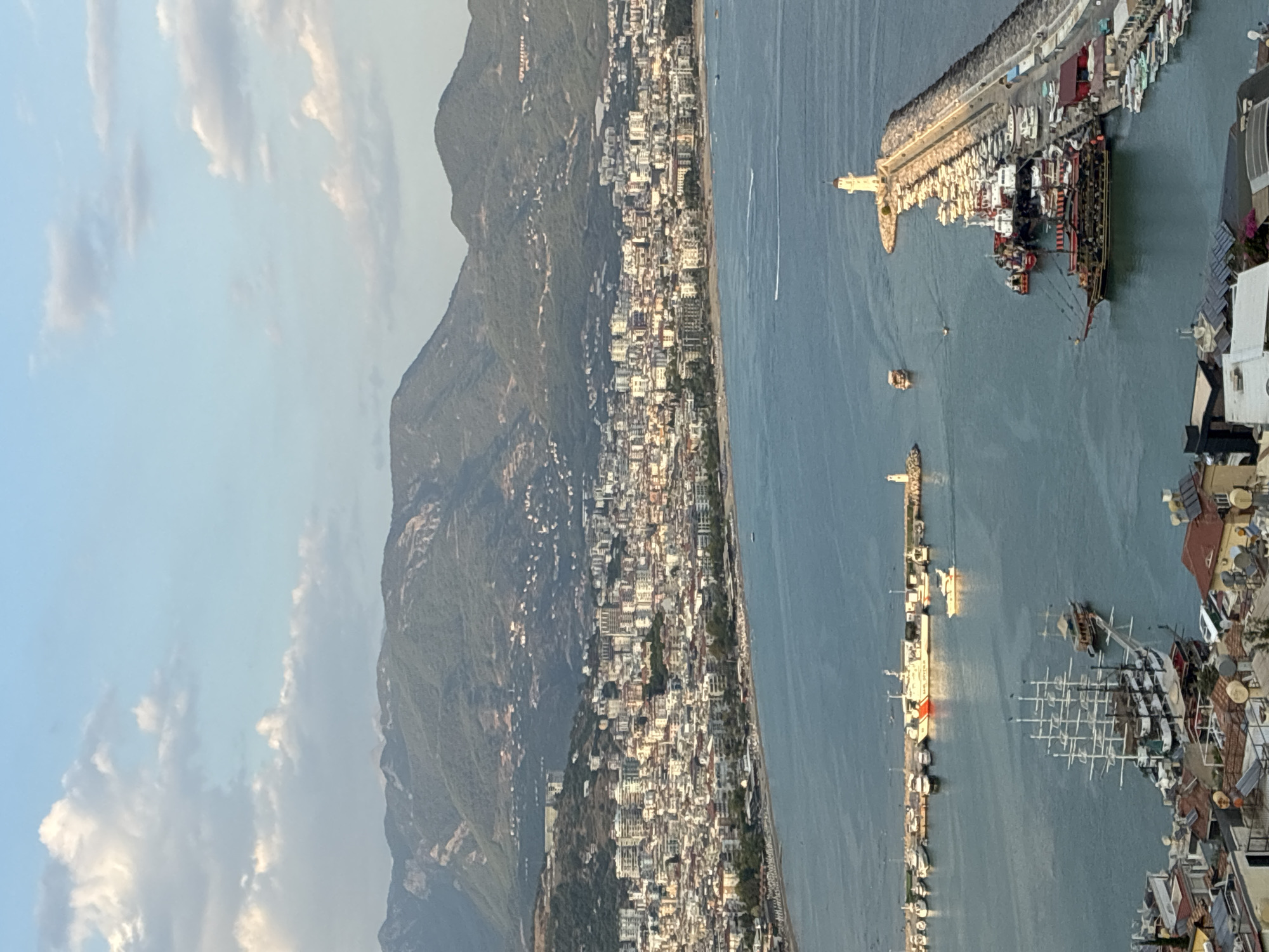 This is a high-angle, medium-shot photograph of the harbor and coastline of Alanya, Turkey, as seen from a higher elevation. The scene is dominated by the bright blue water of the Mediterranean Sea, which stretches from the foreground to the middle ground. Along the coastline, a densely populated city with white, beige, and red-roofed buildings climbs up the slopes of a large, verdant mountain range in the background. The mountain range is covered in a mix of trees and scrub, with some buildings and roads visible on its lower slopes.
In the foreground and middle ground, the harbor is filled with a variety of boats. A prominent breakwater extends into the sea on the right side of the image, lined with numerous docked pleasure boats and yachts. At the end of the breakwater stands a white lighthouse. Further into the harbor, a large, multi-masted sailing ship, likely a tourist vessel, is moored. Several smaller boats, including speedboats and a large, dark-colored pirate-themed ship, are also visible. A coast guard vessel with "GULF OF ANTALYA" written on its side is docked along the breakwater.
The water is relatively calm, with some gentle ripples and white wakes from moving boats. A few people are visible on the beach in front of the city, and small groups are scattered on some of the boats. The sky above is a pale blue, with scattered white, fluffy clouds that catch the light. The overall lighting suggests it is late afternoon or early evening, with a soft, warm glow over the scene.