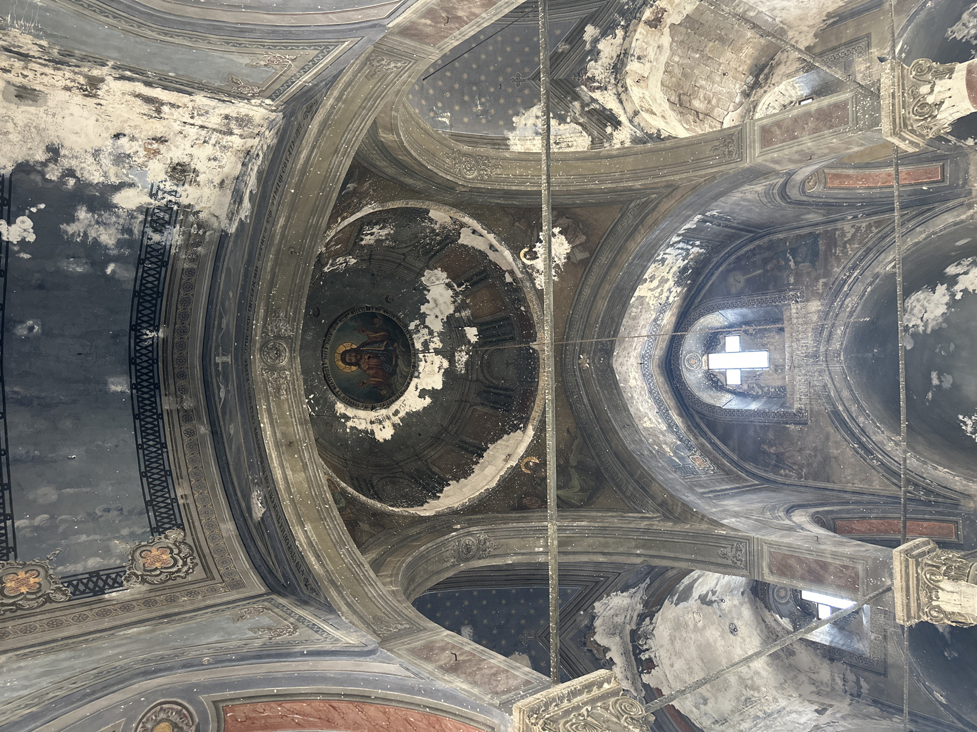 The image depicts the interior of a grand, aged church in Derinkuyu İlçe Merkezi, Turkey. The architecture features high, arched ceilings adorned with faded frescoes and intricate decorative patterns. A prominent dome in the center displays a circular depiction of Jesus Christ, surrounded by celestial motifs. Evidence of decay is visible throughout, with peeling paint, water stains, and crumbling plaster on the walls and ceiling. Sunlight streams through arched windows, illuminating the dusty interior and casting shadows across the vast space. The overall impression is one of historical significance and profound abandonment.