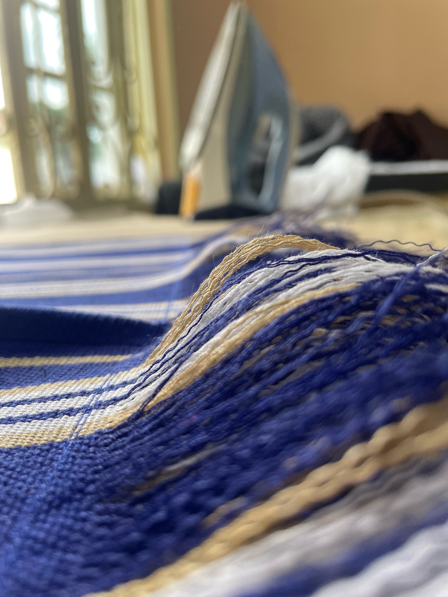 The image is a close-up, low-angle shot of a striped textile, possibly a rug or fabric, with prominent blue, white, and beige stripes. The textile features a fringed edge composed of loose threads. In the blurred background, an iron is visible, suggesting the textile might be in the process of being ironed. The environment appears to be indoors, with natural light streaming in from what looks like a window with curtains. The overall scene is static and domestic. The location in Gura Top, Nigeria, is not discernible from the visual information present in the image.