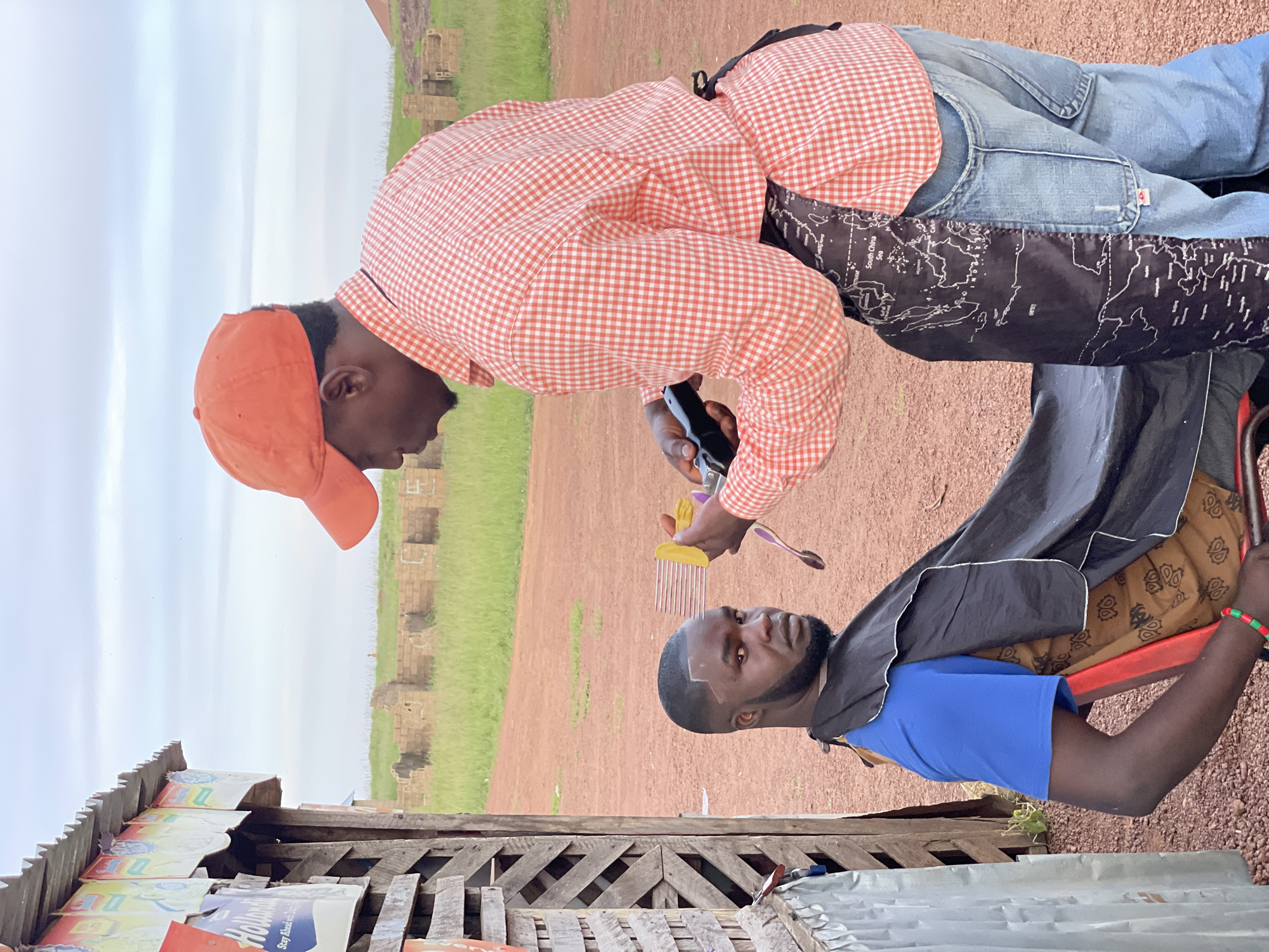 Two men are the subjects of this photograph. One man, wearing an orange checkered shirt, an orange cap, and blue overalls with a map design, is in the process of giving the other man a haircut. He holds a comb and scissors in his hands. The man receiving the haircut, wearing a blue t-shirt and a dark cape, has a distinctive geometric haircut. The scene appears to be outdoors, with a dirt ground and a grassy field with some brick structures in the background. The sky is overcast, suggesting a cloudy day. In the foreground, there is a wooden structure with various signs, one of which has partially visible text. The overall mood of the image seems to be casual, possibly capturing a moment of daily life or a service being provided in a rural setting. There is a bracelet visible on the arm of the man receiving the haircut, which has green, red, and yellow beads.