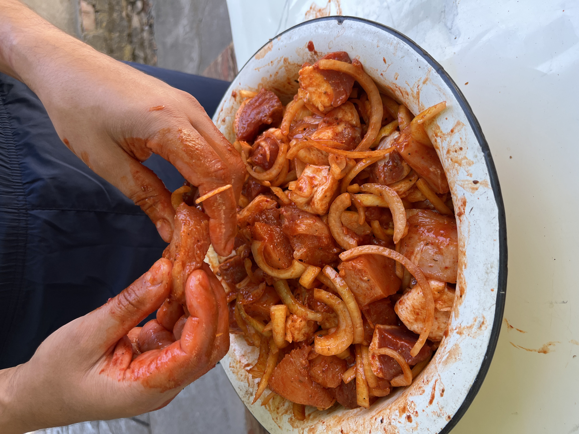 A close-up, overhead shot captures a pair of hands, coated in a red marinade, gently arranging pieces of marinated meat and sliced onions in a large, white enamel bowl. The bowl rests on a light-colored surface, possibly a table, with a hint of a gray, brick wall and outdoor foliage visible in the background, suggesting an outdoor setting. The marinade is a vibrant red-orange color and liberally coats the food items and the hands. The hands are positioned above the bowl, actively manipulating the contents. The bowl itself is speckled with remnants of the marinade, indicating it has been used for mixing. The overall scene suggests the preparation of a meal, likely for grilling or marinating. The specific location in Taraz city administration, Kazakhstan, cannot be determined from the visual information provided in the image.