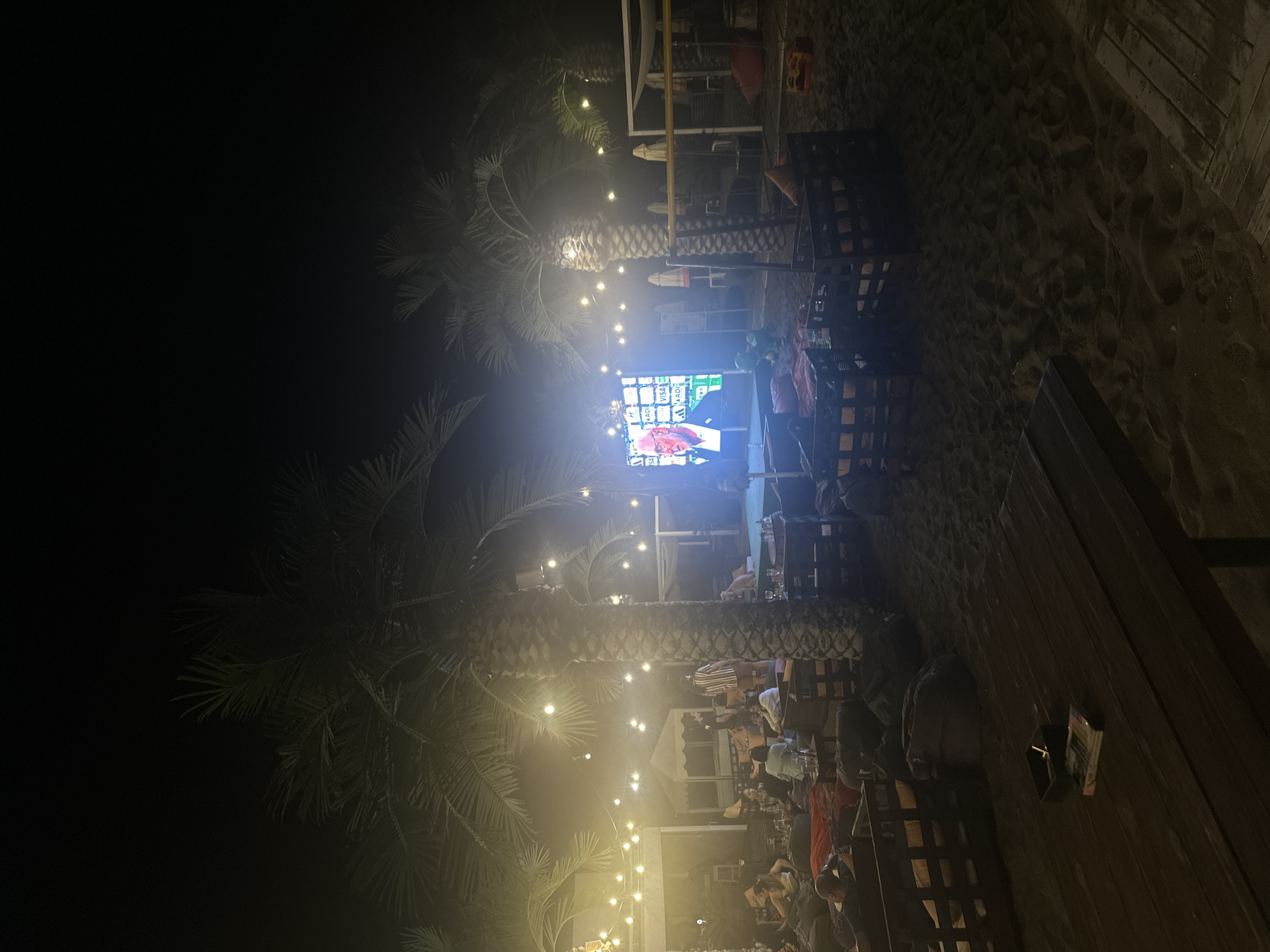The image depicts an outdoor night scene, likely at a beachside establishment in Burgas, Bulgaria. Several people are visible, seated on beanbag chairs and around low tables on a sandy surface. A large LED screen is prominently displayed, showing a close-up of a man's face, suggesting an event like a sports broadcast or a presentation is taking place. Palm trees with string lights draped between them create an ambient atmosphere. In the foreground, a wooden table holds an ashtray and a pack of cigarettes. In the background, beach umbrellas and other seating arrangements are visible, along with a volleyball net. The overall impression is of a relaxed gathering or event taking place after dark.