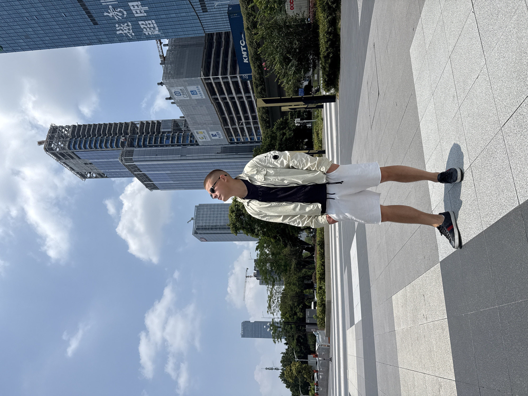 A young man stands facing left, positioned on a wide, light-grey tiled pavement under a bright sky in an urban environment, identified as Huangpu District, China. He appears to be in his twenties, wearing a light cream-colored bomber jacket over a black t-shirt, white shorts, and dark athletic shoes with red and white accents. He has short hair and sunglasses, obscuring his expression.
The setting features modern high-rise buildings, several with glass facades, dominating the background. One prominent building is under active construction, with scaffolding and exposed structural elements. Another building features large Chinese characters on its side, including "粤海" (Guangdong Sea/Ocean) and characters associated with "建筑" (construction). Further back, there are more completed skyscrapers and green spaces with trees and shrubs. A large blue shipping container, bearing the text "KMTCL.jpn" and "OOCL," is visible among the greenery and construction.
The time of day is sunny, indicated by clear skies with scattered white clouds and strong, defined shadows cast by the buildings and the man onto the pavement, suggesting midday or early afternoon. The man is standing still, observing his surroundings. The scene depicts a bustling, modern city undergoing development.