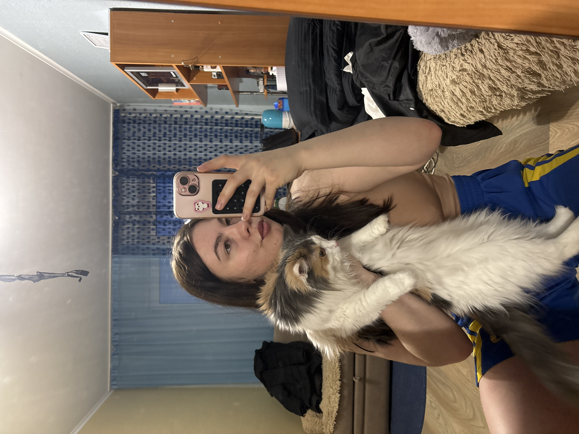 A woman is holding a long-haired cat while taking a selfie in front of a mirror. The woman is wearing blue athletic shorts with yellow stripes. The background shows a bedroom with a wardrobe, blue patterned curtains, and bedding. The exact location in Perm, Russia cannot be determined with the given information.
