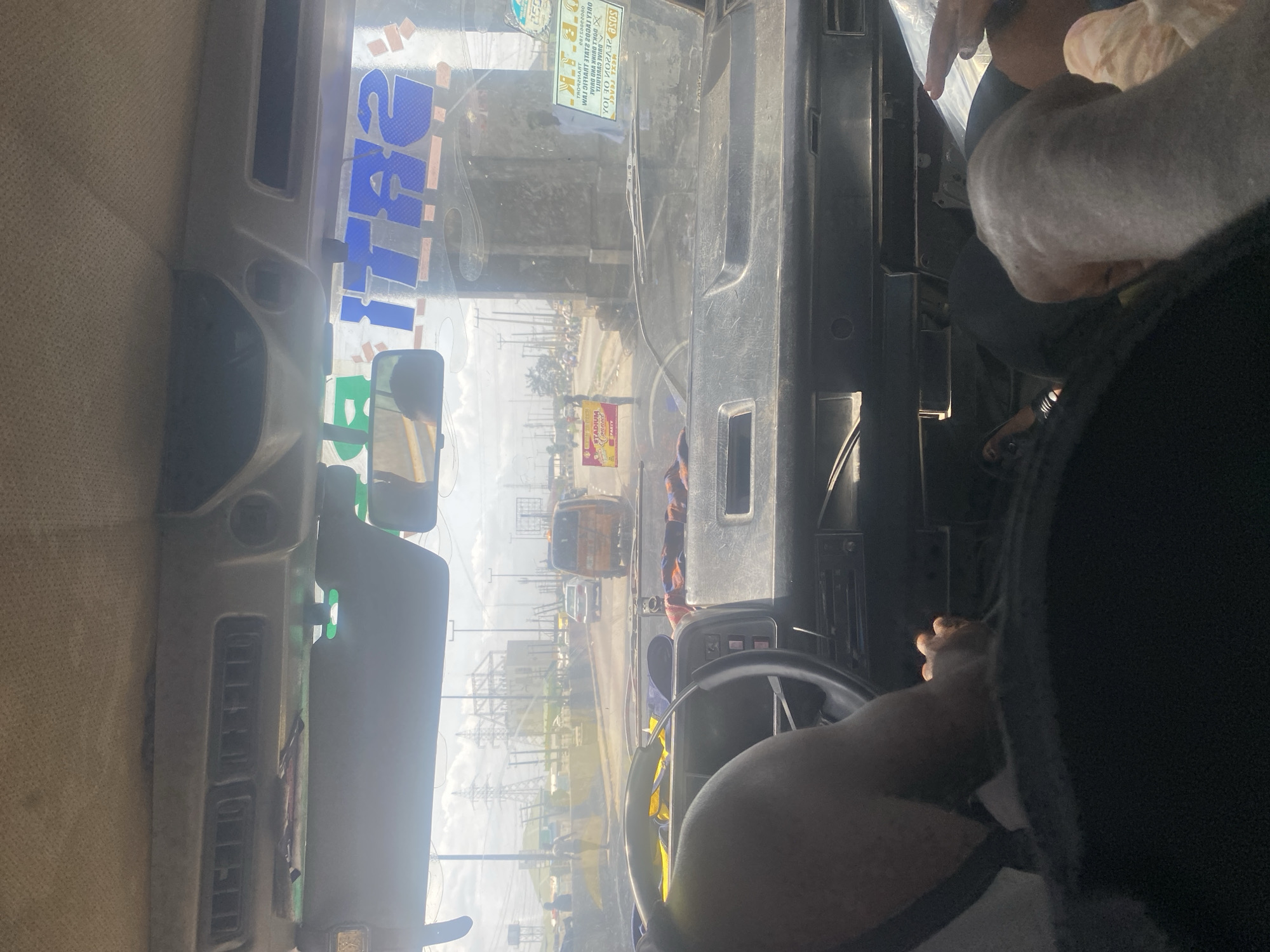 The image shows the interior of a vehicle, likely a bus, from the driver's perspective. The driver's hands are on the steering wheel. The view through the windshield reveals a street in Unknown, Unknown. There are utility poles, buildings, and vehicles visible on the street. A yellow sign, possibly an advertisement, is visible on the street. The OCR suggests the presence of a stadium.