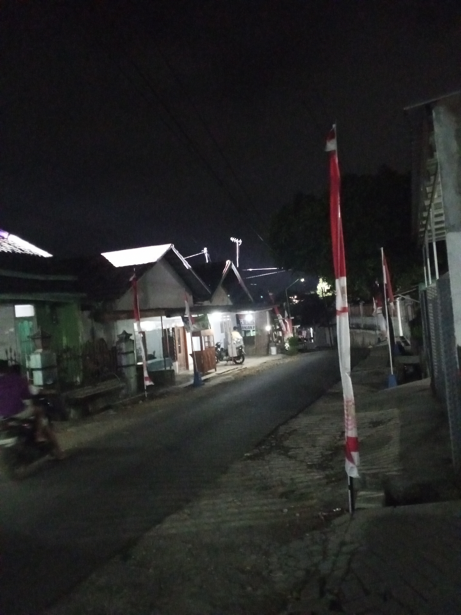 The image depicts a street scene at night in Turen, Indonesia. The street is paved and runs between a row of buildings on the left and a sidewalk with a building on the right. Several red and white flags are prominently displayed on poles along the street. The buildings on the left appear to be houses or small businesses, some with lit windows. A motorbike is visible on the left side of the street, with a person seated on it, illuminated by a purple light. On the right side, a tall, slender object with red and white banding extends upwards, likely a flag pole. The sky is dark, indicating nighttime. The overall impression is of a quiet, possibly residential or commercial street decorated for an event.