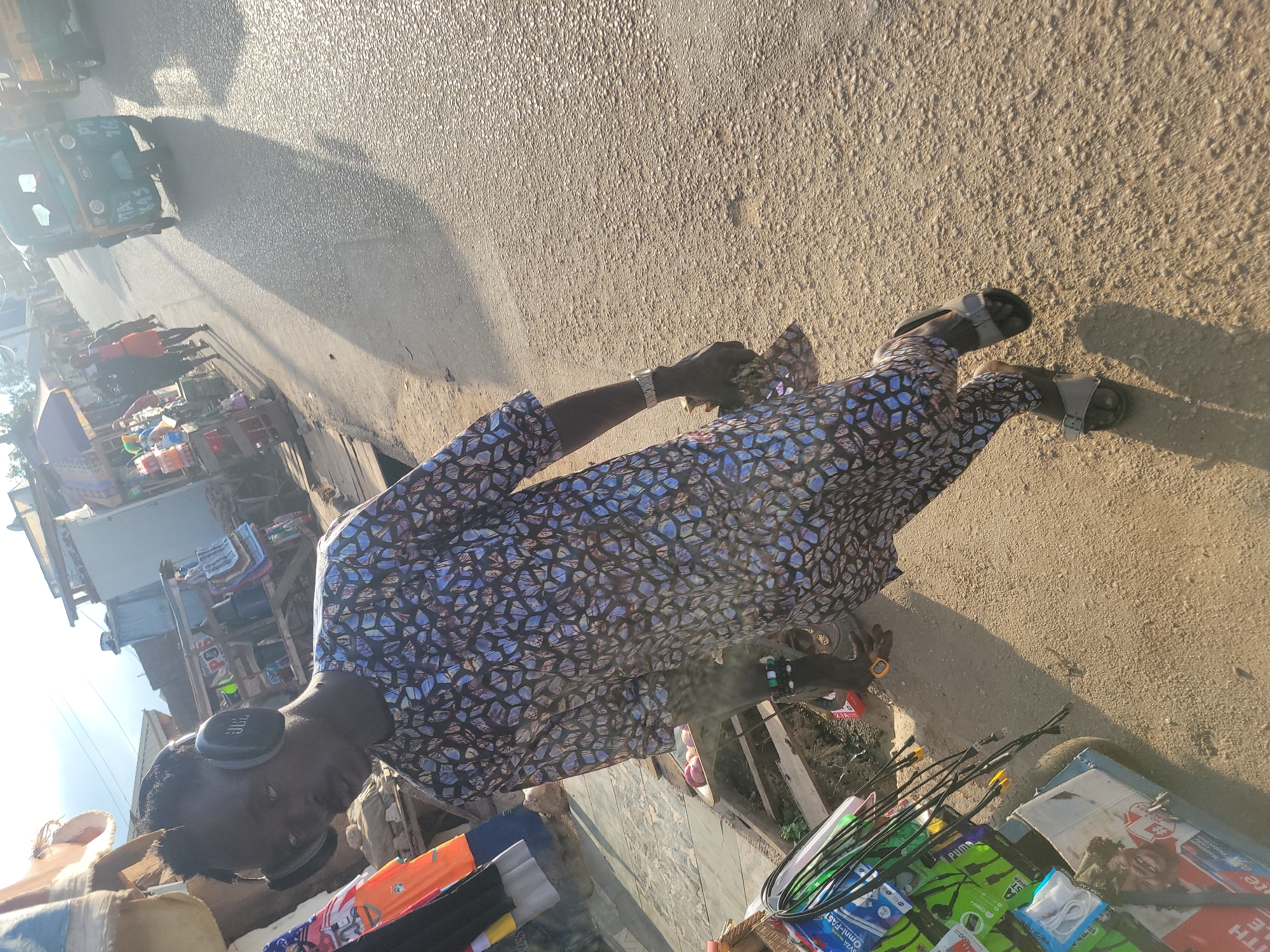 The image depicts a bustling street scene in Jos, Nigeria, likely an urban market area, illuminated by strong, low-angle sunlight, indicating either early morning or late afternoon.
In the foreground, a male individual, appearing to be a young adult, with a dark complexion and short dark hair, is walking towards the viewer's left. He is wearing a long-sleeved, patterned tunic (kaftan-style) and matching trousers, featuring a blue, black, and white geometric or abstract design. Over his ears, he wears black JBL brand over-ear headphones. A silver-colored watch is visible on his left wrist, and a multi-colored beaded bracelet adorns his right wrist. He carries a small, indistinct item in his right hand and is wearing grey sandals. He casts a prominent long shadow extending behind him and to the left.
To the immediate left of the main subject, a makeshift stall displays various merchandise, primarily electronic accessories such as charging cables, headphones, and packaged goods. Brands like "Omni-Fast," "Puma," and "Tecno" are partially visible on packaging, alongside a "Colgate" box featuring a child's image. The stall is constructed from wood and appears to be part of a larger market setup.
The ground is a mix of asphalt and dirt, showing textured surfaces due to the sunlight. In the mid-ground and background, the street stretches into the distance, populated by other pedestrians and several tricycles, commonly known as Keke Napep in Nigeria. One green tricycle in the mid-ground clearly displays "PTA KAS" and "PTA 76" on its front, identifiers common for commercial vehicles in Plateau State, where Jos is the capital. Buildings and additional market stalls line both sides of the street further down, contributing to a vibrant, dense urban environment. The sky above is bright, with some atmospheric haze.