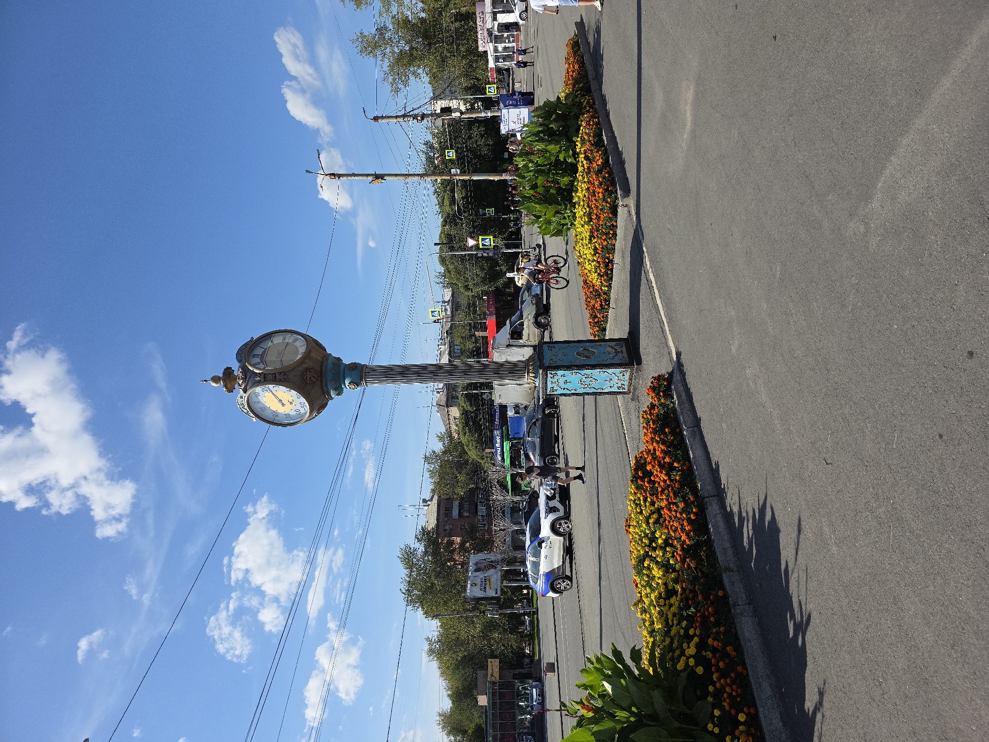 This is a daytime, eye-level, medium shot of a public square in Chelyabinsk, Russia, featuring a large clock tower as the central element. The clock tower, with its ornate, blue and gold design, stands tall on a lamppost-like structure. It has two clock faces, one showing approximately 10:07 and the other around 22:07, suggesting it indicates both local time and perhaps another time zone or has separate day and night indicators. Below the clock faces, there are what appear to be thermometers, one showing readings in Celsius with a range from -50 to +50.
The square is paved, with a road visible in the foreground and middle ground. The road is populated by various vehicles, including a white car with blue markings, a black car, a white van, and what appears to be a bus in the background. People are present, with one person walking across the street and another riding a bicycle. Pedestrian crossings with yellow warning signs are visible.
The environment is characterized by a clear blue sky with scattered white clouds. Lush green trees line the background, interspersed with buildings that suggest an urban setting. In the foreground, there are vibrant flower beds filled with orange and yellow flowers, adding a splash of color to the scene. The overall impression is of a lively urban public space on a sunny day.