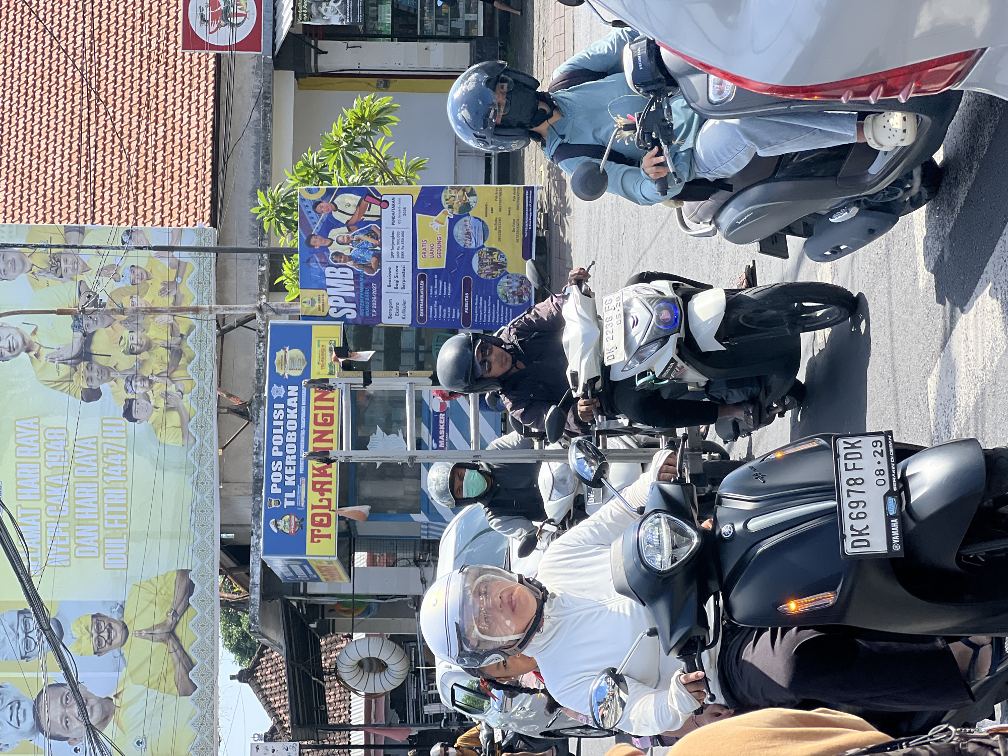 The image shows a busy street scene in North Kuta, Indonesia. Several people are riding motorbikes in close proximity. The closest rider wears a white helmet and jacket. The plate number on the bike nearest the camera reads DK 6978 FDK, with an expiration sticker for 08.29. Other visible plate numbers read DK 2238 FC. Buildings line the street, adorned with large banners and signage. Banners for local events are visible on the roadside. An advertisement for a school named SPMB and a poster featuring local leaders are displayed, as well as a sign for a police post called "POS POLISI TL KEROBOKAN".
