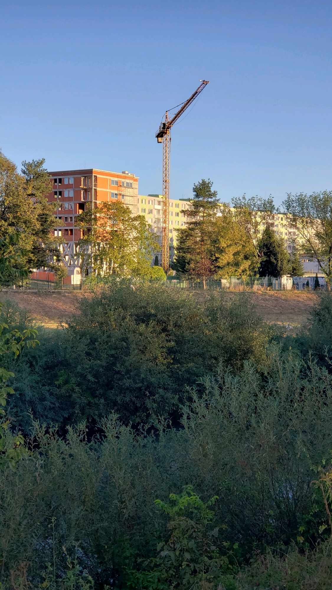 The image depicts a construction site in Prešov, Slovakia, with a tall yellow tower crane dominating the center of the frame. The crane is positioned in front of several multi-story residential buildings, some of which appear to be under construction, indicated by exposed brickwork and scaffolding on one building. The buildings are a mix of orange and light yellow facades. Lush green trees and foliage frame the lower portion of the image, creating a contrast between nature and urban development. The sky is a clear, bright blue, suggesting a sunny day. The scene appears to be a suburban or residential area undergoing development.