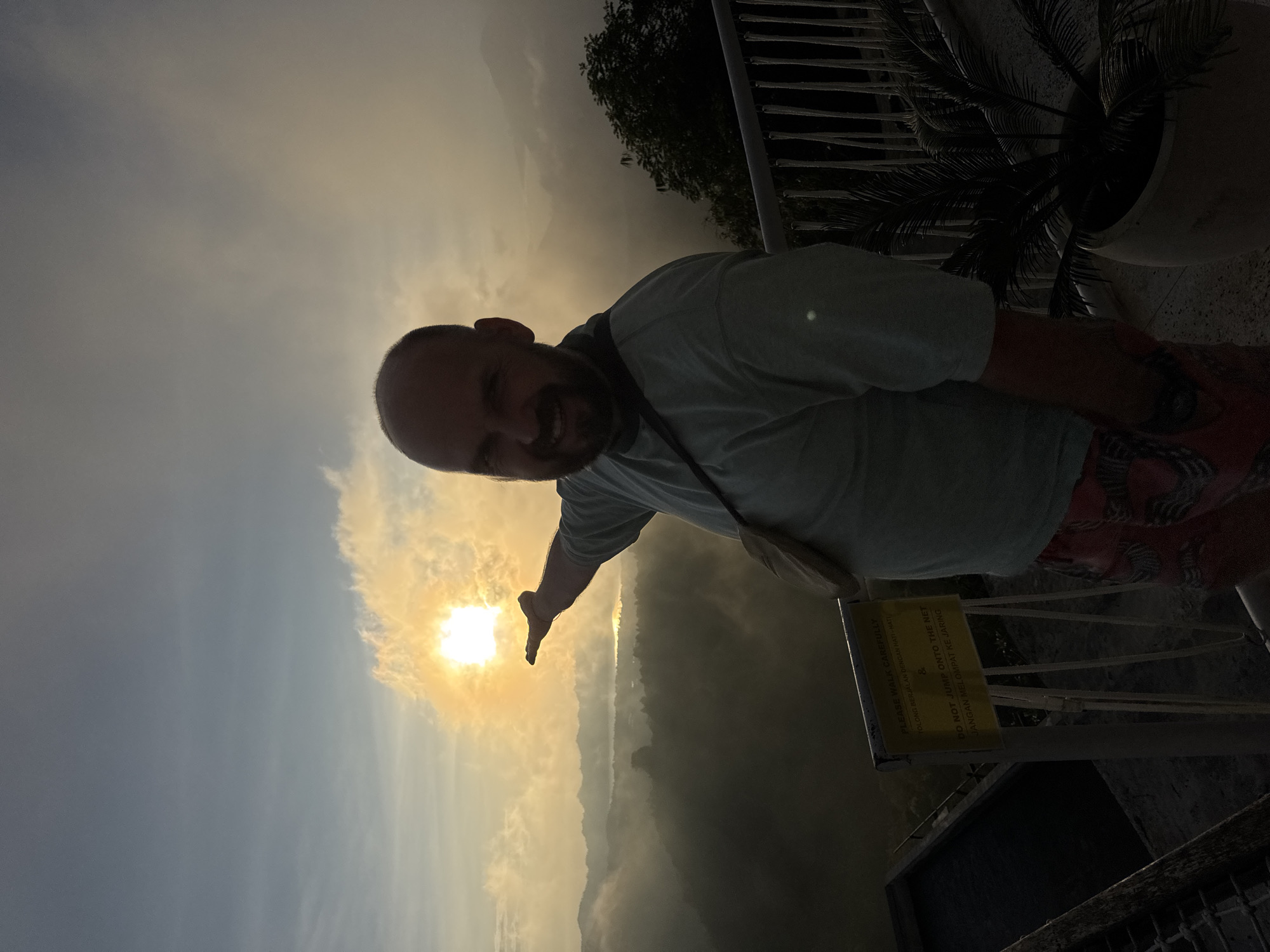 The image depicts a man with a beard and short hair, likely in his 30s or 40s, standing on a ledge and reaching out towards the sun, seemingly holding it in his hand. He is wearing a light-colored t-shirt and patterned shorts. He has a bag slung over his shoulder.
The scene appears to be at a high vantage point, possibly a cliff or observation deck, overlooking a vast landscape shrouded in mist or fog. A sign written in English and Indonesian warns visitors to walk carefully and not to jump onto the net, indicating a protective barrier nearby.
The location is likely in Kedisan, Indonesia, based on the Indonesian language on the sign. The sky is bright with the sun shining through a thin layer of clouds. The overall impression is one of a tourist enjoying a scenic view at sunrise or sunset. There are plants and railings visible, suggesting a constructed viewing area.