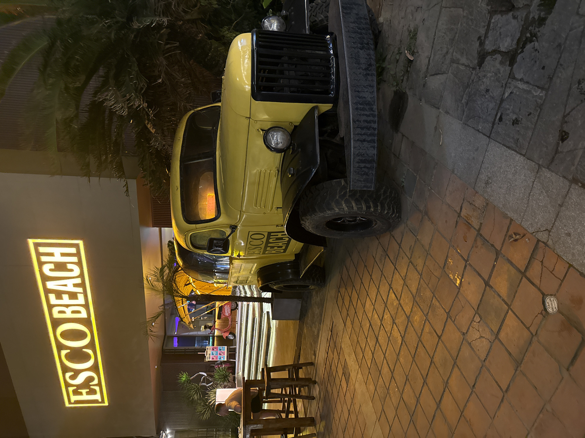 The image depicts a yellow, vintage military-style vehicle parked outside the ESCO BEACH bar and restaurant in Đà Nẵng, Vietnam. The vehicle has a black grille and bumper, and the name of the establishment is painted on its side. The ground is paved with light orange, square tiles. A seating area with wooden tables and chairs is located near the vehicle. The ESCO BEACH sign is illuminated with bright yellow lights. The scene appears to be at night. There is one person at a table.