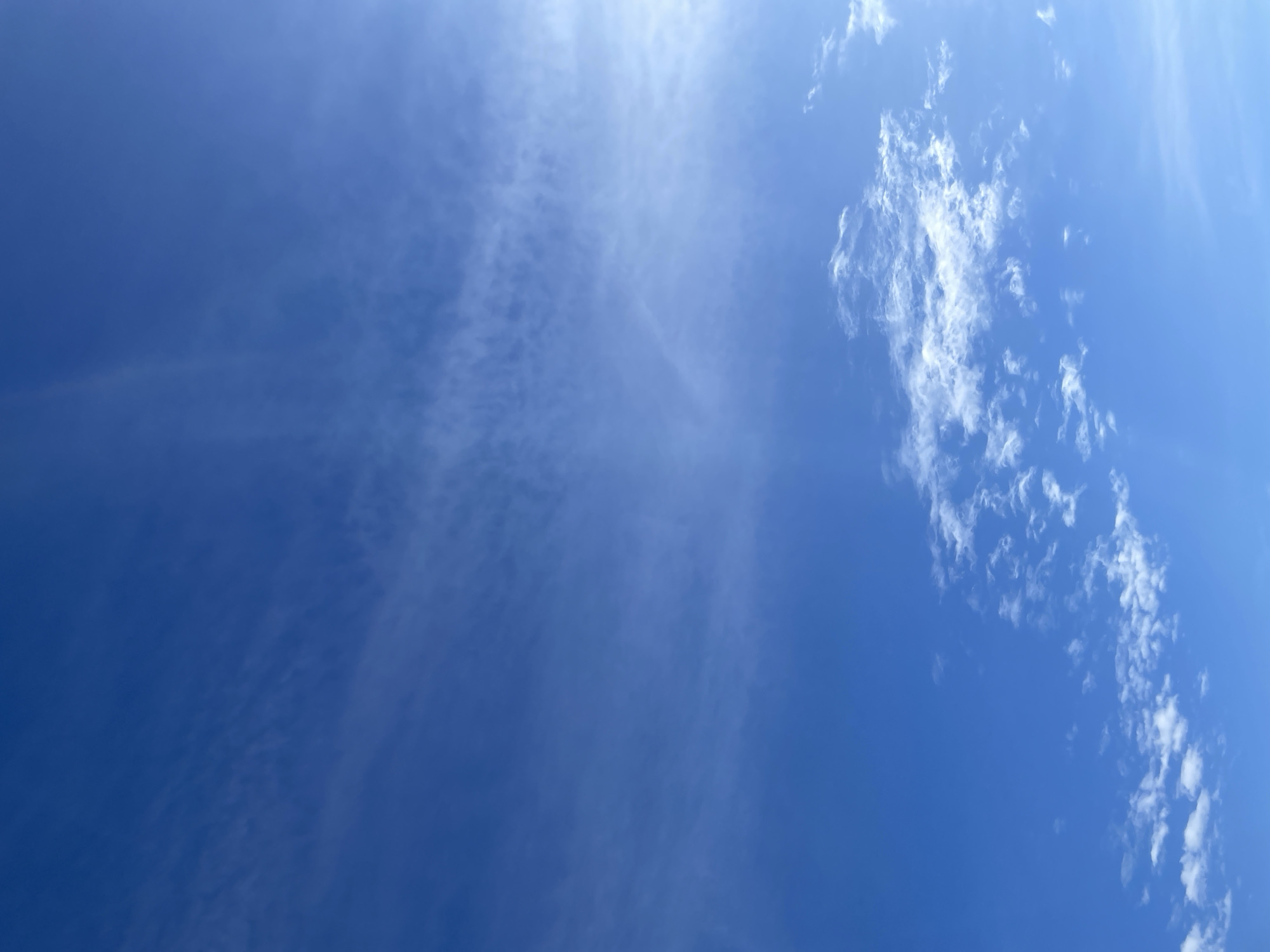 The image displays a blue sky with varying cloud formations, observed over the city of Zav'yalovo, Russia. The sky exhibits a gradient of blue, transitioning from a deeper hue on the left to a brighter, sun-illuminated area on the upper right. Thin, wispy, semi-transparent clouds, consistent with cirrus or cirrocumulus types, are distributed across the upper and central portions of the frame. In the lower right quadrant, several smaller, more distinct white cloud fragments, resembling fragmented cumulus or stratocumulus, are visible. No terrestrial features, structures, or individuals are present within the captured scene.