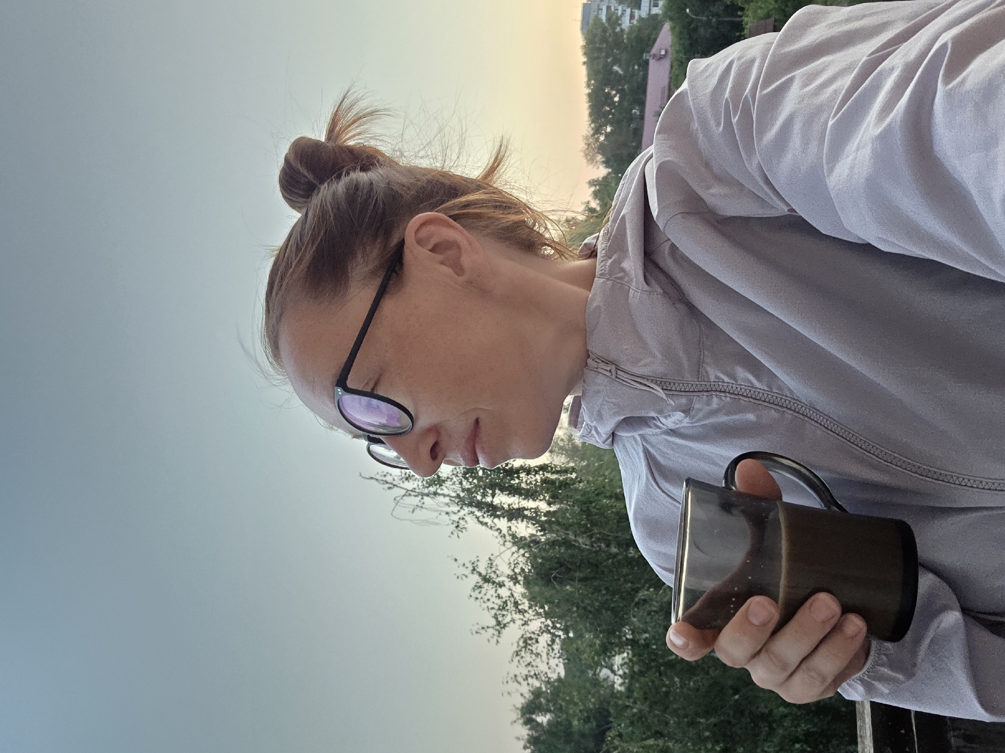 A woman with blonde hair pulled into a bun and wearing glasses and a light-colored jacket is holding a mug of coffee. She is looking to her left, out at a view of trees and buildings. The sky is clear and pale blue, suggesting it is either early morning or late evening. The scene appears to be set on a balcony or elevated vantage point, with a railing visible in the foreground. The location is identified as Chaikovsky, Russia.
