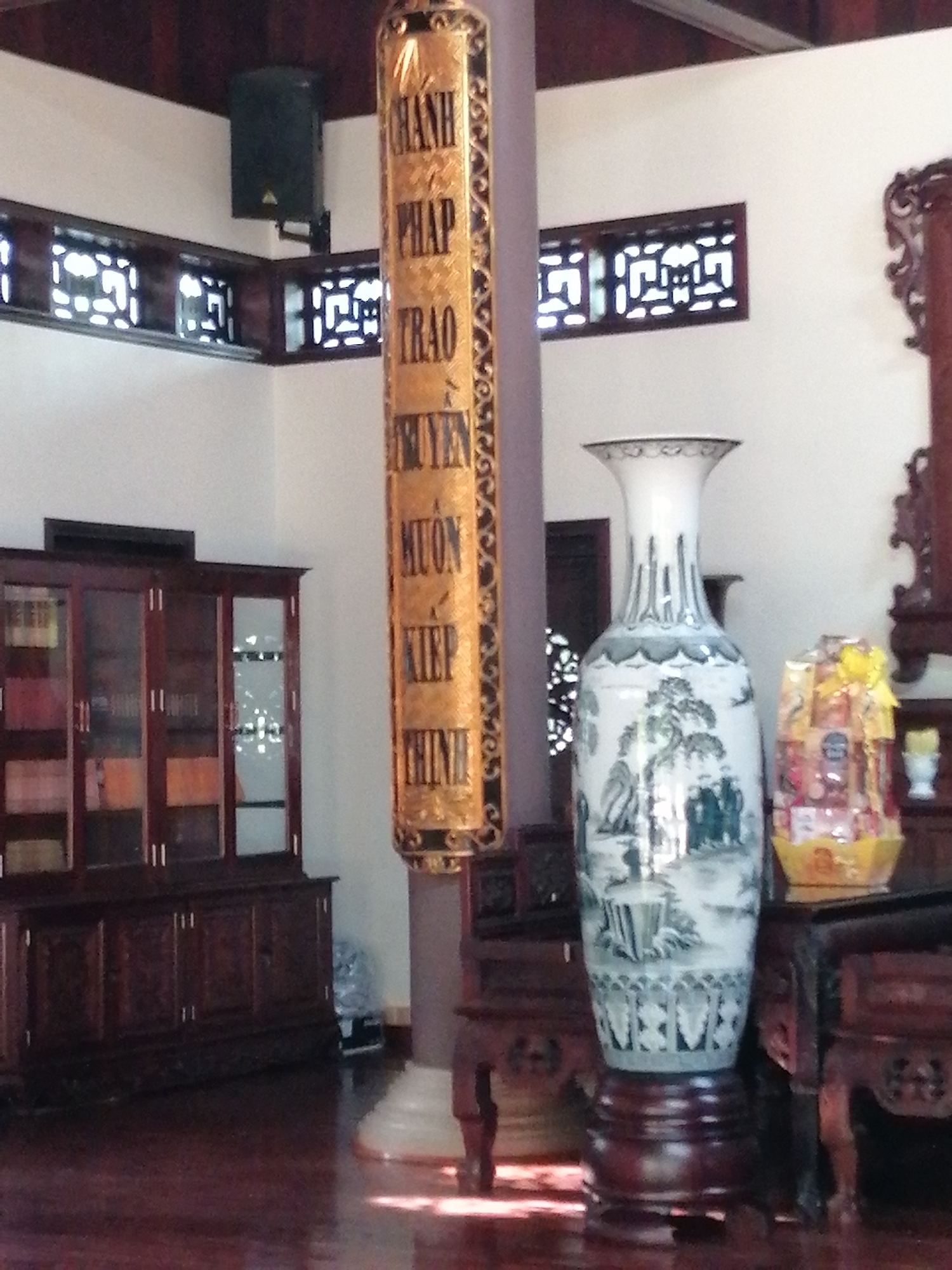 The image captures an interior scene with traditional Vietnamese decor. A large, ornate golden pillar with inscribed Vietnamese text stands prominently in the foreground, leaning slightly to the right. To the right of the pillar, a tall, white ceramic vase with blue intricate designs sits on a dark wooden stand. Next to the vase is a gift basket filled with various items, wrapped in yellow cellophane. Behind the vase and gift basket, a dark wooden cabinet with glass doors is visible, displaying various items within. In the upper left of the image, a black speaker is mounted on the white wall, with a decorative wooden lattice window above it. Another similar lattice window is seen further to the right, above the cabinet. The floor is made of dark polished wood, reflecting the light. The overall impression is one of a well-decorated, possibly religious or cultural space, likely within a home or a temple.