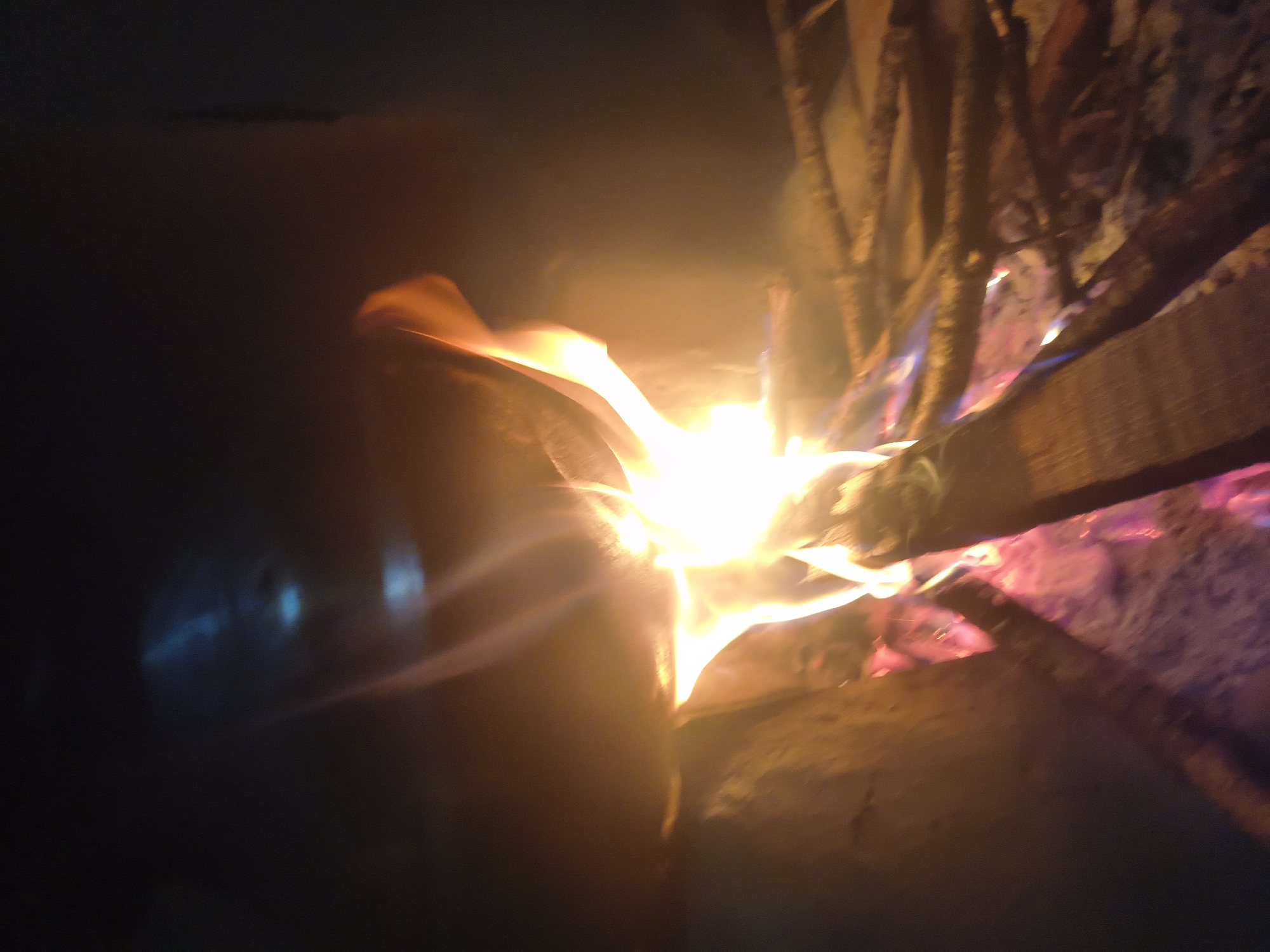 The image captures a close-up view of a bonfire with flames reaching upwards. Several pieces of firewood are visible, some partially consumed by the fire, while others are stacked or arranged. The background is dark, with subtle hints of light reflections in the upper portion of the image, possibly from a metallic surface. The ground beneath the fire appears to be ash and embers, with tinges of red and purple from the heat. There are no people or animals visible in the frame. The scene suggests an outdoor setting at night, where a fire is being maintained. There is no specific information in the image to indicate the location within Haini, India.