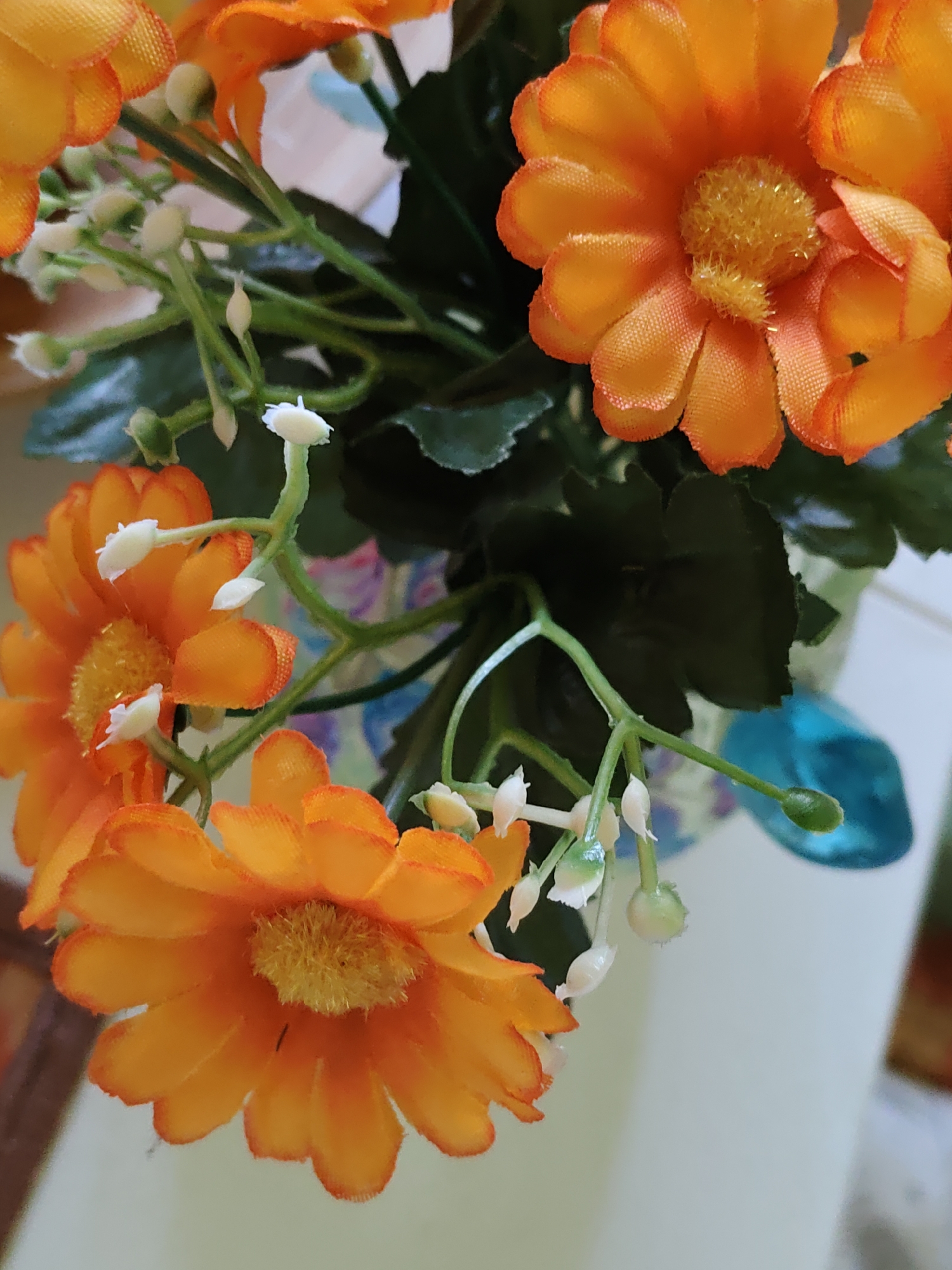 The image shows a close-up of a bouquet of artificial orange flowers with yellow centers, adorned with small white buds. The flowers are arranged on green stems and leaves. In the background, a blue decorative object, possibly a vase or a part of a planter, is visible. The setting appears to be indoors, with a soft, neutral-colored background. The image was taken in Rostov-on-Don, Russia.