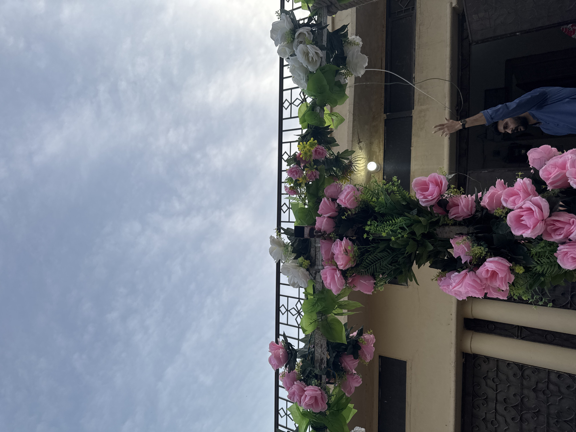 The image shows a building facade in Justice Colony, Pakistan, decorated with a floral arrangement of pink and white roses cascading down a black iron railing. A man in a blue shirt extends his arm from a window on the right side of the frame. The sky is overcast with scattered clouds. The building is painted in neutral tones. There are windows with dark frames.