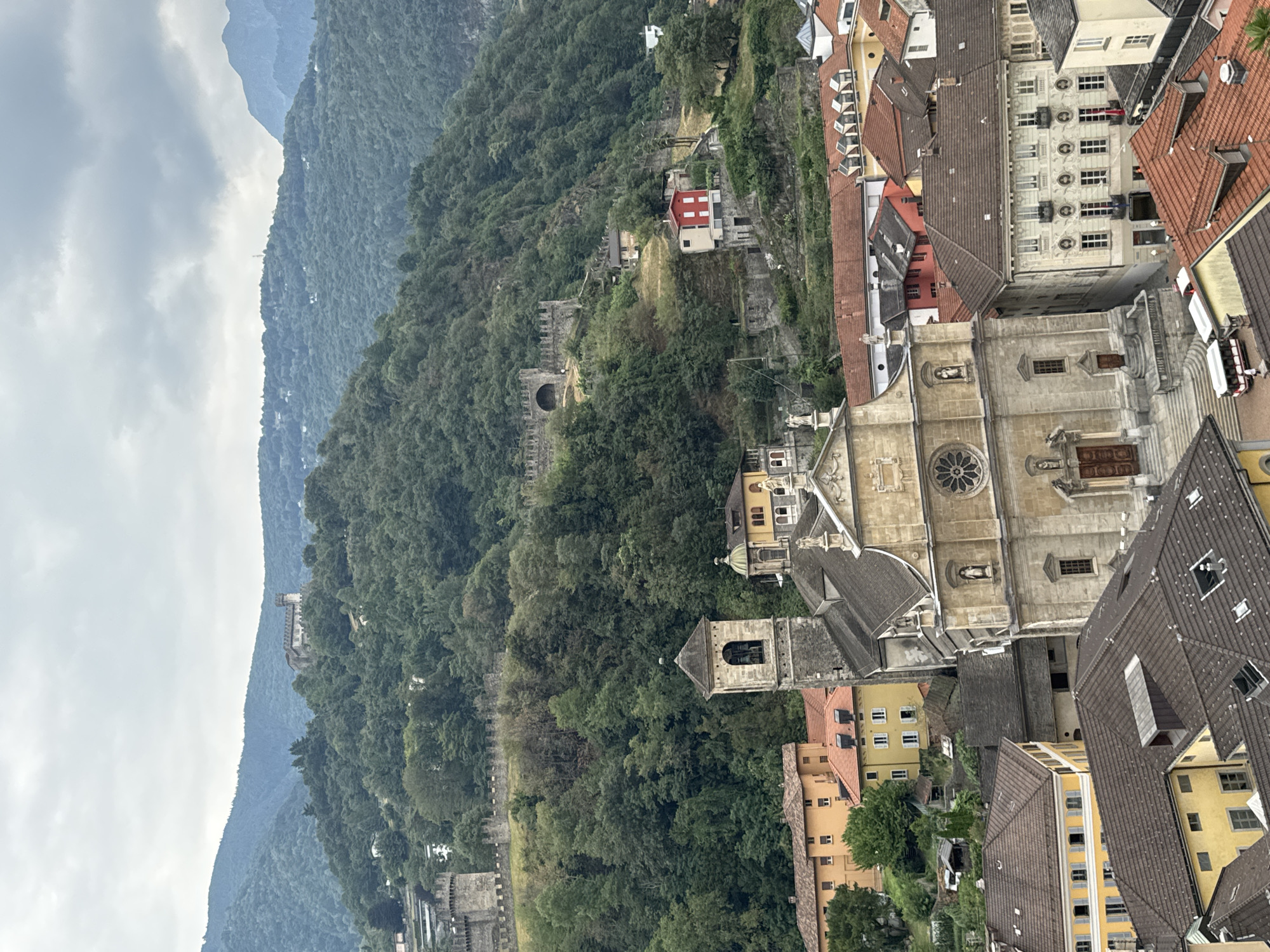 The image depicts a panoramic view of the city of Bellinzona, Switzerland, featuring the distinctive UNESCO World Heritage Castles of Bellinzona. In the foreground, the ornate facade of a church with a prominent bell tower is visible, surrounded by terracotta-tiled roofs of surrounding buildings. Behind the church, the ancient fortifications of Castel Grande and Montebello stretch across the verdant, forested hillside, culminating in a distant, more isolated castle structure atop the highest peak. The sky is overcast, casting a muted light on the scene. The overall impression is one of historical depth and natural beauty, with the medieval castles serving as a striking counterpoint to the urban and natural landscape.