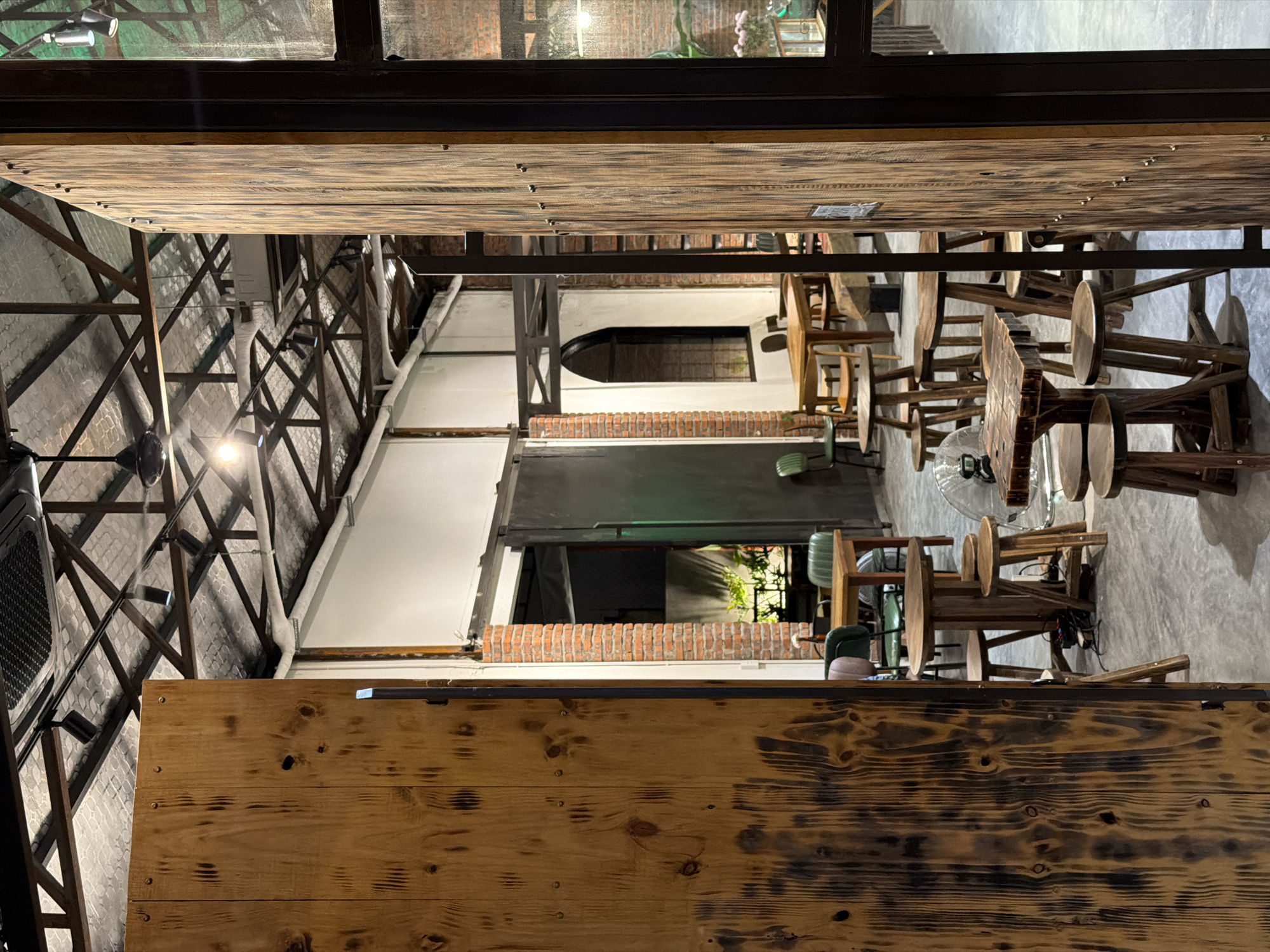 The image depicts an interior view of a building in Đà Nẵng, Vietnam. The structure features a high, partially visible ceiling supported by a network of metal beams. A wooden plank serves as a visual barrier in the foreground. Further in the background, there's a dining area with wooden tables and stools. A doorway leads to an outdoor area, revealing greenery. The interior lighting is a mix of natural and artificial sources. The walls are a combination of brick and white plaster. The floors appear to be concrete. The perspective is unusual, possibly taken from a high vantage point or with the image rotated.