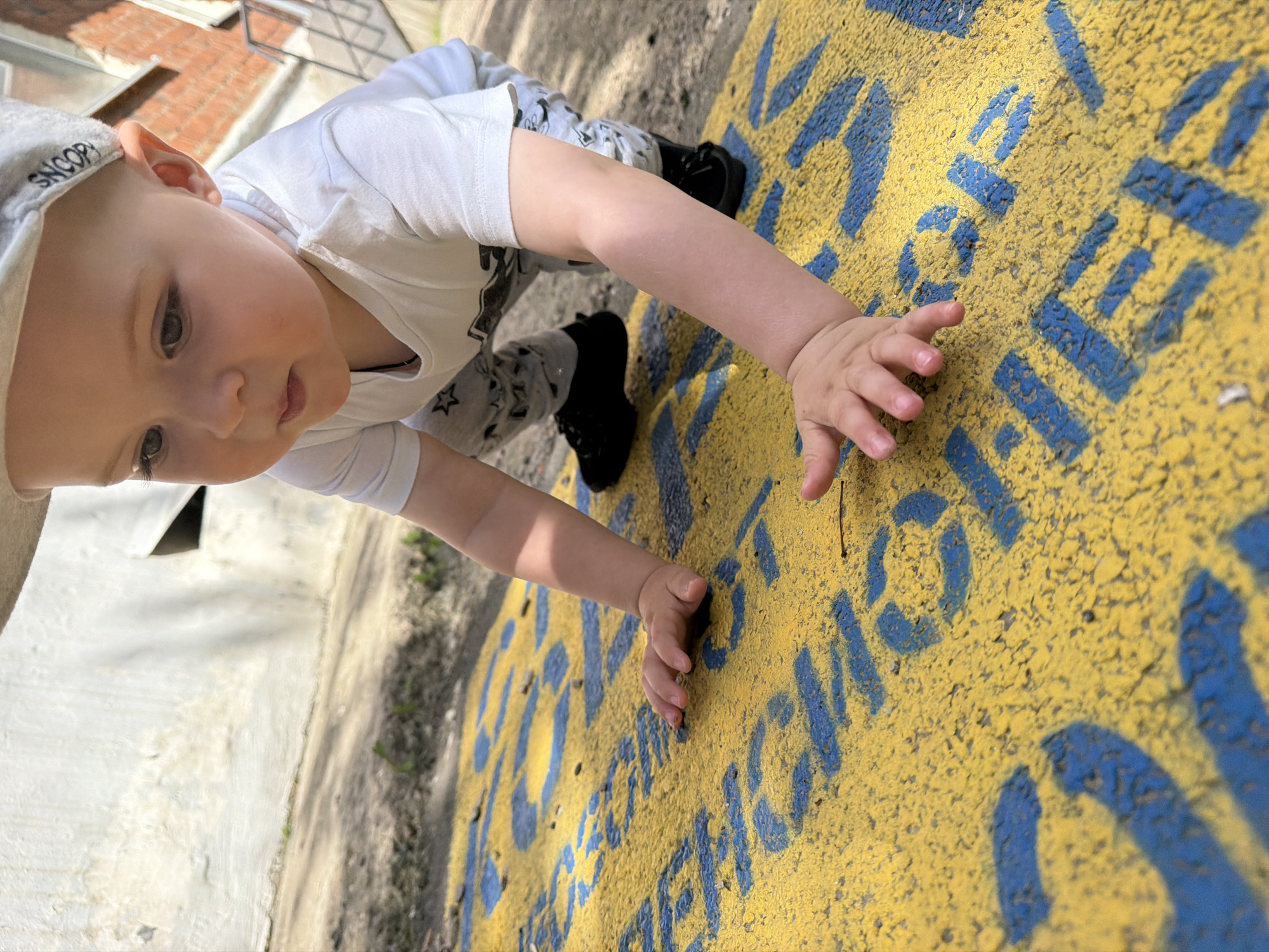 A young child, appearing to be an infant or toddler, with fair skin and light features, is positioned in a crouching or crawling stance. The child wears a light-colored baseball cap with "SNOOPY" visible on the side, a white short-sleeved t-shirt, and grey trousers patterned with black stars. Black shoes are visible on the child's feet. Both of the child's hands are placed on a ground surface that is textured and painted in a worn yellow color, featuring stenciled blue letters and abstract shapes. The child's gaze is directed downwards towards this patterned ground. In the background, a light-colored, textured wall of a building is visible to the left. To the right, part of a reddish-brown brick building and a dark metal railing are present. The scene is illuminated by direct sunlight, casting shadows. This outdoor scene is located in the city of Votkinsk, Russia.