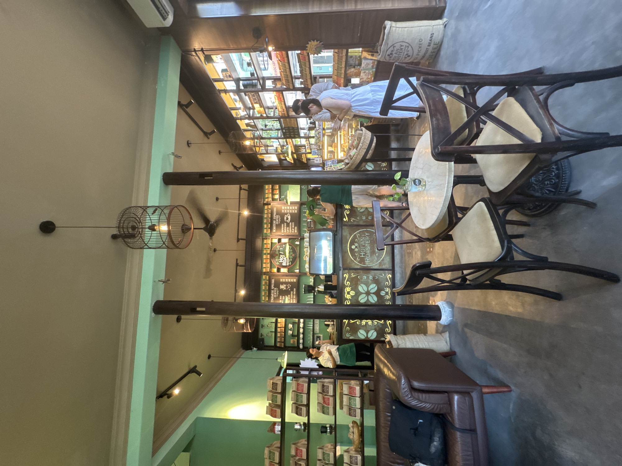 The image depicts the interior of the Hoi An Roastery, a coffee shop in Hoi An, Vietnam. The scene is filled with warm lighting and displays of coffee products. In the foreground, there are several dark wooden chairs with light-colored cushions arranged around a round marble-topped table. To the left, a brown leather sofa is visible.
The middle ground shows a coffee counter with a large espresso machine and various pastries displayed in a refrigerated case. Behind the counter, shelves are filled with bags of coffee. A woman in a green apron is standing at the counter, appearing to be serving a customer. Further back, on the right side of the image, more shelves display coffee products. Two people, a man in a white shirt and a woman in a white dress, are standing at the counter, seemingly interacting with the staff or browsing products.
The environment is characterized by wooden pillars, shelves, and decorative elements. A prominent feature is a birdcage-like light fixture hanging from the ceiling. The walls are painted in a muted green color, and the ceiling has exposed beams and lighting fixtures. The floor appears to be concrete. The overall impression is that of a well-stocked and inviting coffee shop.