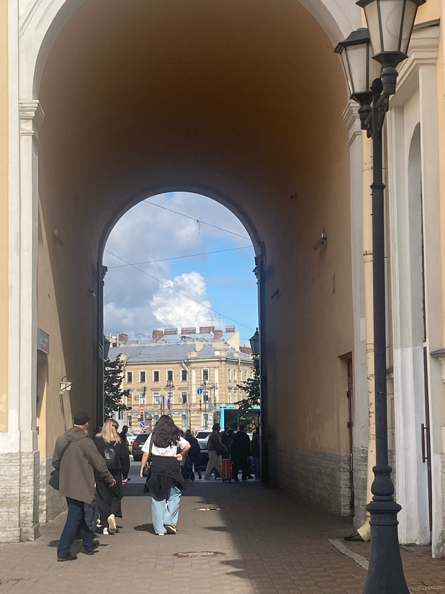The image captures a street scene in Saint Petersburg, Russia, viewed through a large architectural archway. The arch is a prominent feature, casting a shadow across the paved walkway. On the right side of the arch, a black lamppost with two lamps stands tall.
Through the archway, a view of buildings and the sky is visible. The buildings appear to be typical of Saint Petersburg architecture, with light-colored facades and mansard roofs. A bright blue sky with fluffy white clouds suggests a pleasant day. Power lines are strung across the view.
Several people are walking along the paved path. In the foreground, a person wearing a dark hooded jacket and blue jeans walks away from the camera. Next to them are two other individuals, one wearing black clothing and the other in light blue jeans and a white top. Further in the background, more people are seen walking, some with luggage. Cars and a blue bus are visible on the street beyond the archway. The overall impression is of an urban environment with pedestrian activity.