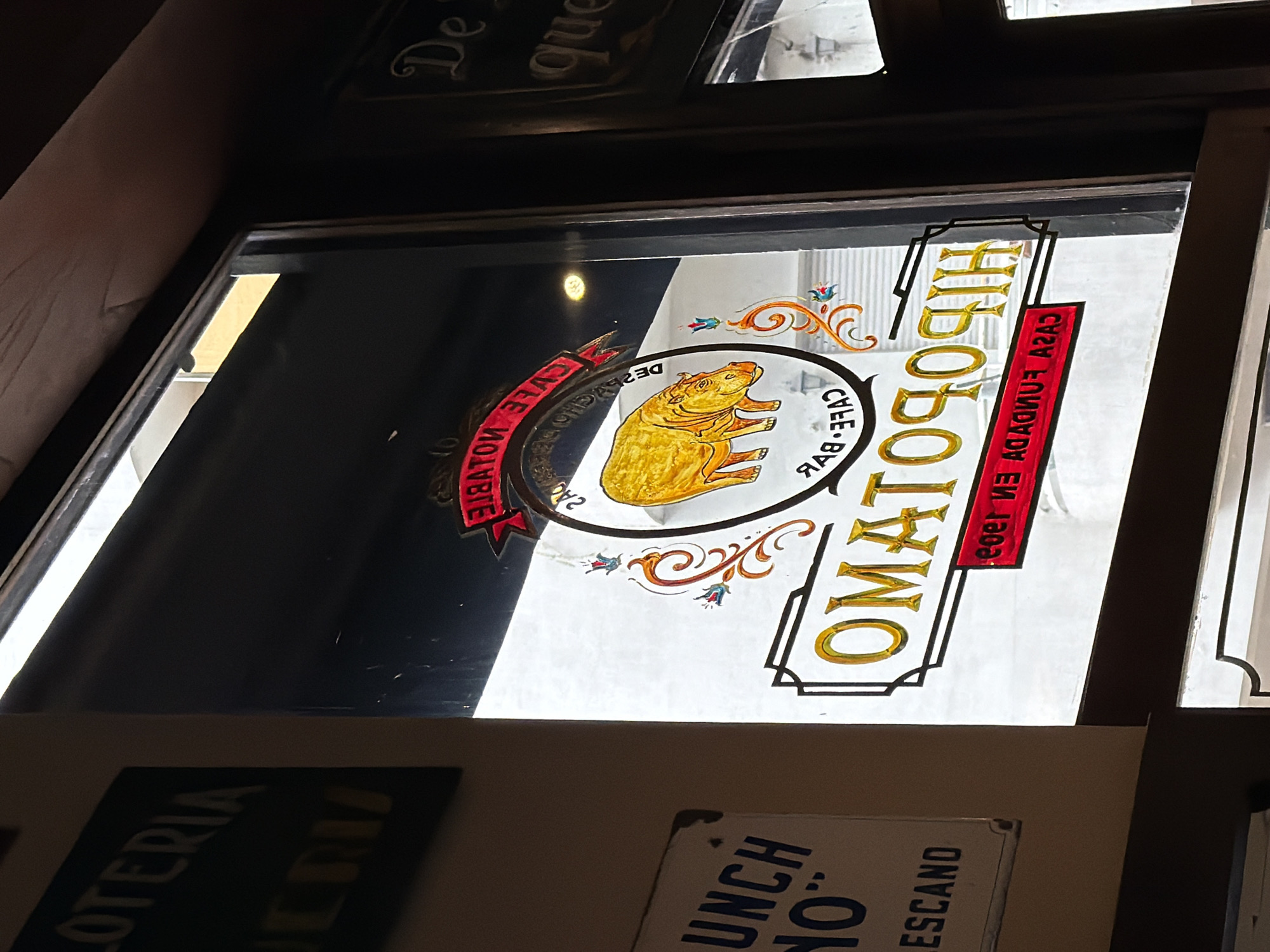 The image shows a vintage sign for "El Homatoche," a "Cafe-Bar" in Buenos Aires, Argentina. The sign is displayed in a glass window, likely of the establishment itself. The sign features a cartoon capybara, and text in Spanish: "CASA FUNUADA EN 1903" (ESTABLISHED IN 1903). Partial signs can be seen below the window: "Lotería" and "Lunch No."