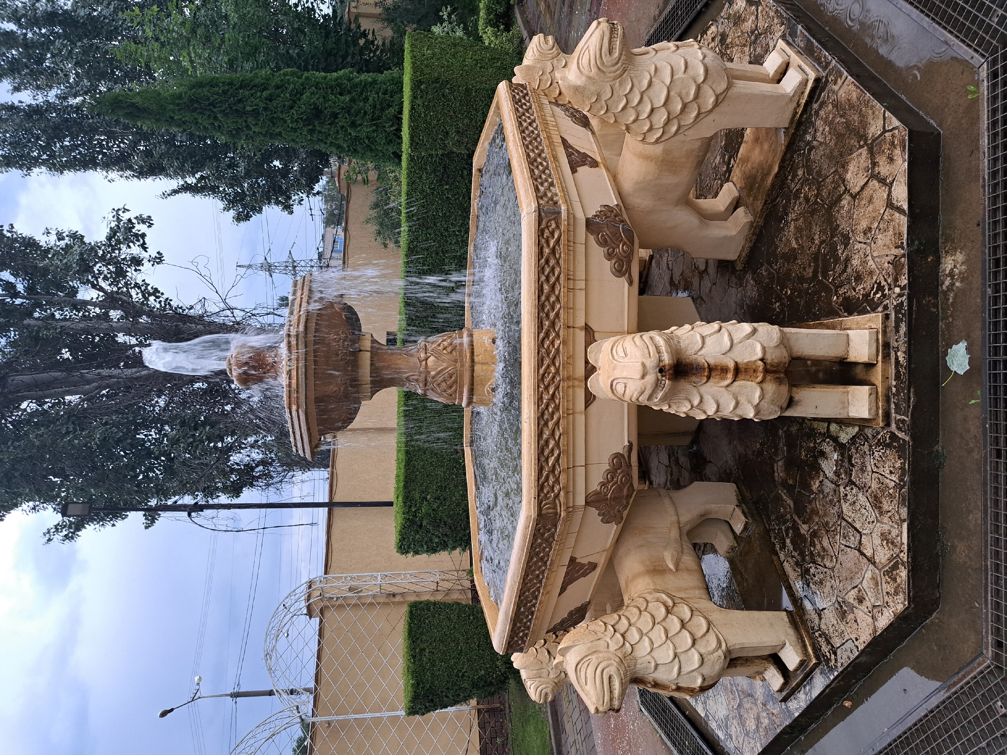 A fountain with lion sculptures at its base is the main subject of this outdoor scene. The fountain is made of a light-colored material, possibly stone or concrete, and features multiple tiers with water cascading down. The lion sculptures are stylized, with carved manes and facial features. The fountain is situated on a textured hexagonal base, surrounded by wet pavement. In the background, there are manicured green hedges, trees, a building with a yellow wall, and a white decorative fence. The sky is overcast, with visible power lines and a street light adding to the urban context. The overall impression is one of a decorative garden or courtyard feature.