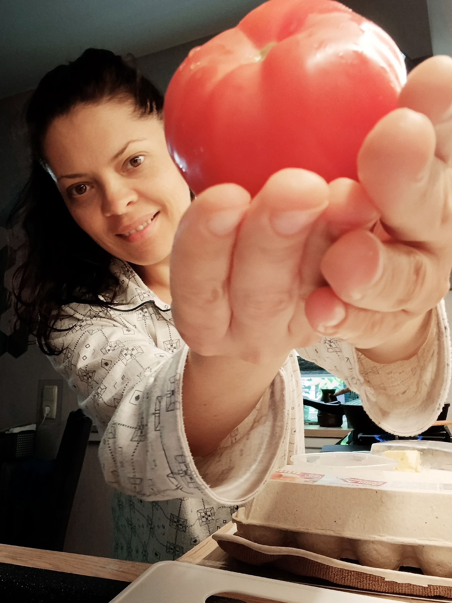 A woman, with dark hair and brown eyes, wearing a light-colored patterned pajama top, is holding a large red tomato towards the camera with her right hand. She is smiling. In the foreground, there is a carton of eggs and a container of butter. The background appears to be a kitchen with a stove and a window. The location is unknown, as there are no visible landmarks or specific details to identify it as Saratov, Russia.