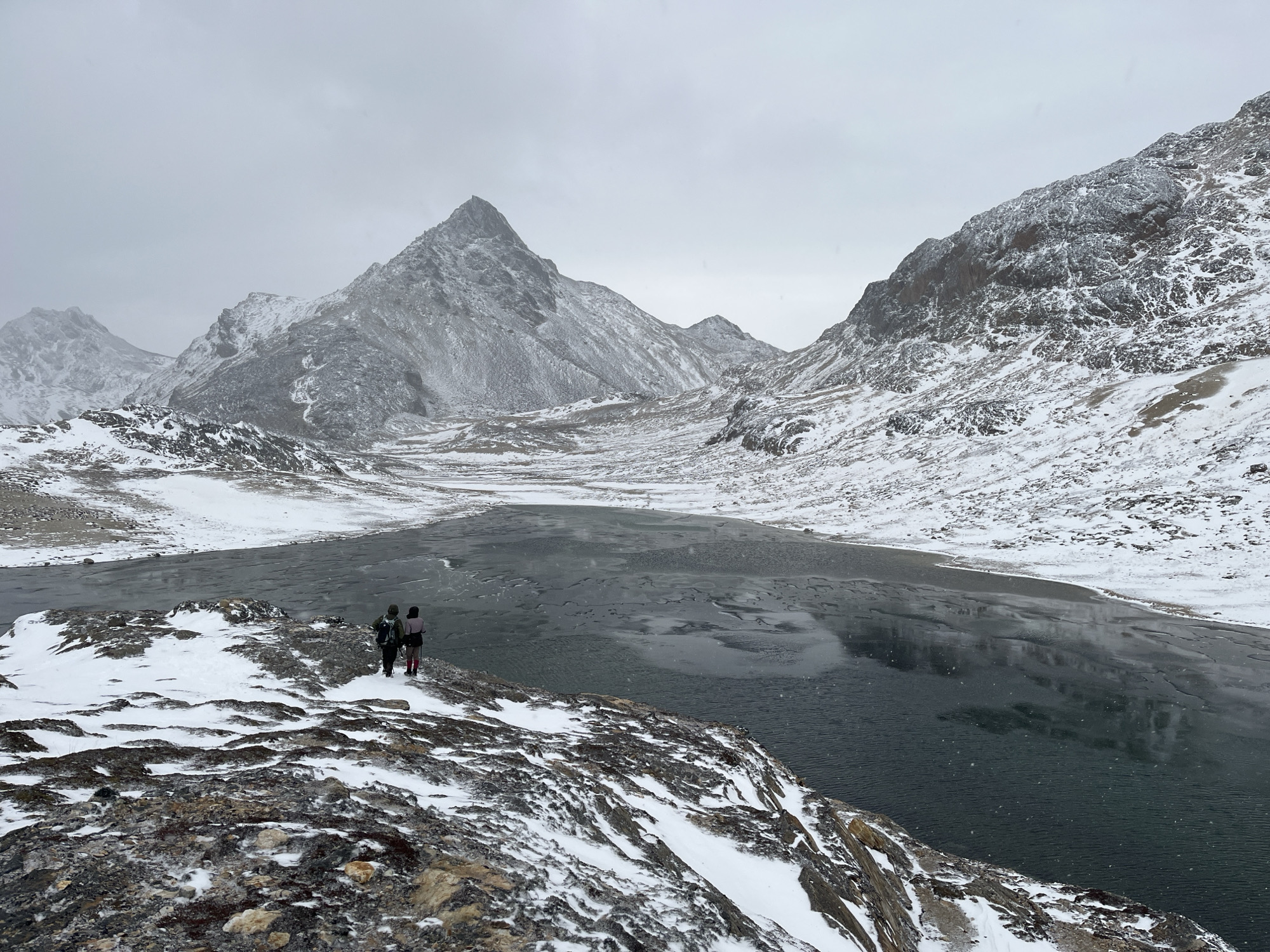 Two people stand on a snowy, rocky outcrop overlooking a dark lake. The background consists of snow-covered mountains under a cloudy sky. The location is not specified, but it's likely somewhere in Argentina given the mention of Ushuaia. Without more information, the exact location in the city cannot be determined.