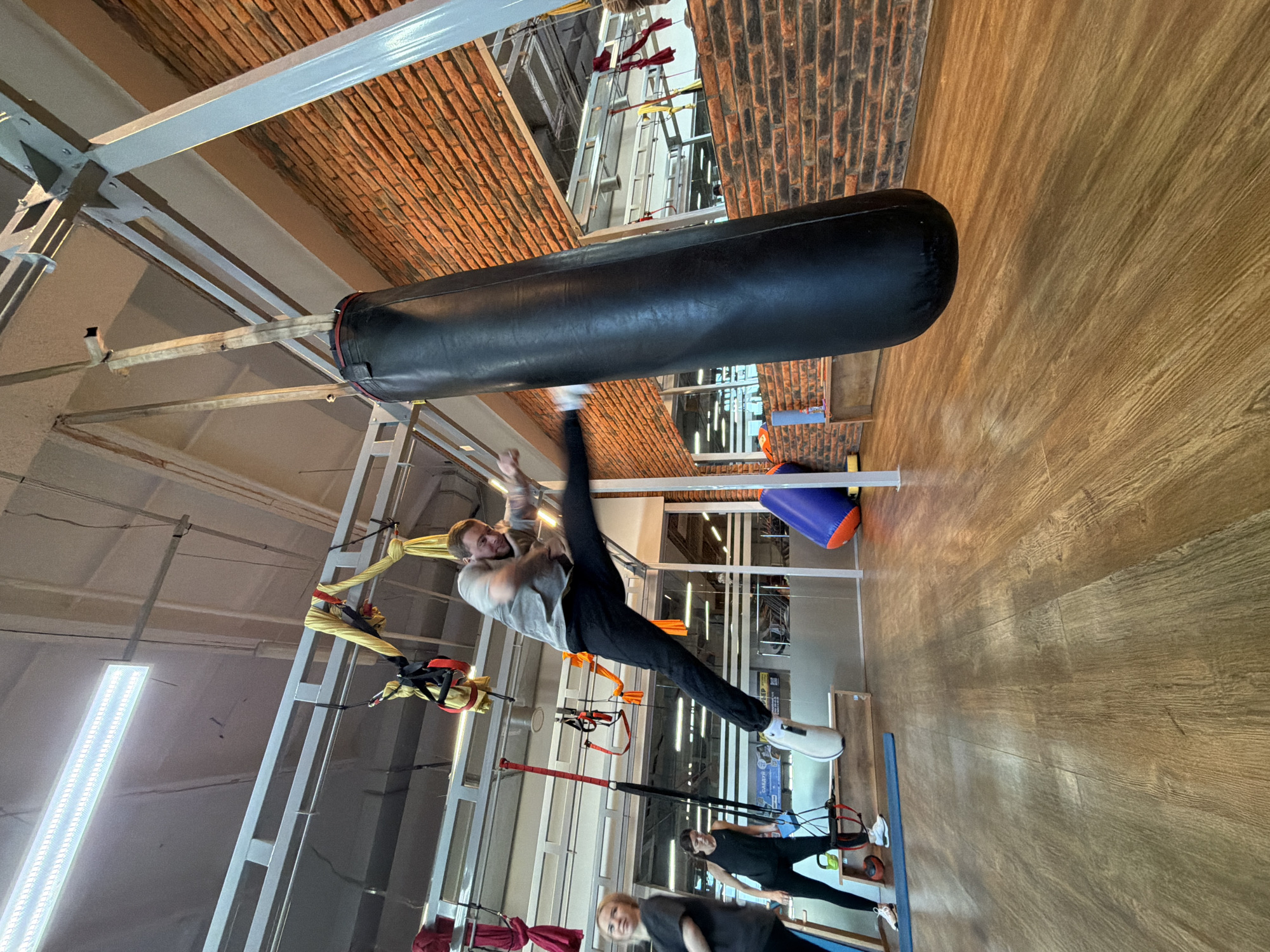 The image shows a man in a gray sweatshirt and black pants performing a high kick towards a black punching bag in an indoor gym setting. The gym has wooden floors and a brick-patterned ceiling. Other equipment includes white metal frames supporting various suspension devices. Two other people can be seen on the floor below near a blue mat. The location is identified as Chaikovsky, Russia.