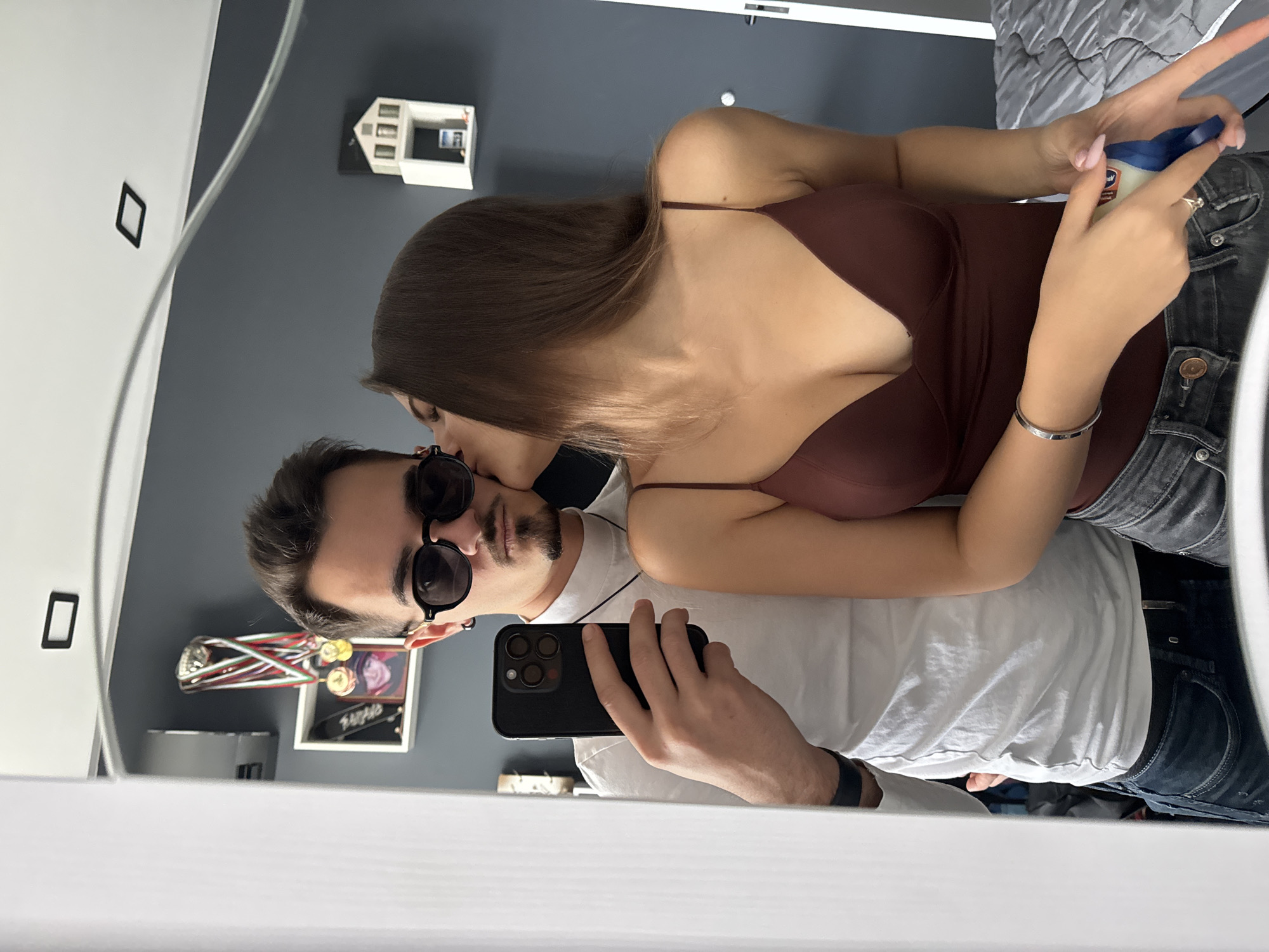 A couple is reflected in a mirror. The woman, with long brown hair, leans over the man, seemingly kissing his cheek. She wears a brown top and jeans and holds a blue and white object. The man wears sunglasses and a white t-shirt, holding a phone. The background suggests a bedroom setting. There is no direct evidence in the image to place the location within Sofia, Bulgaria, or any other specific city.