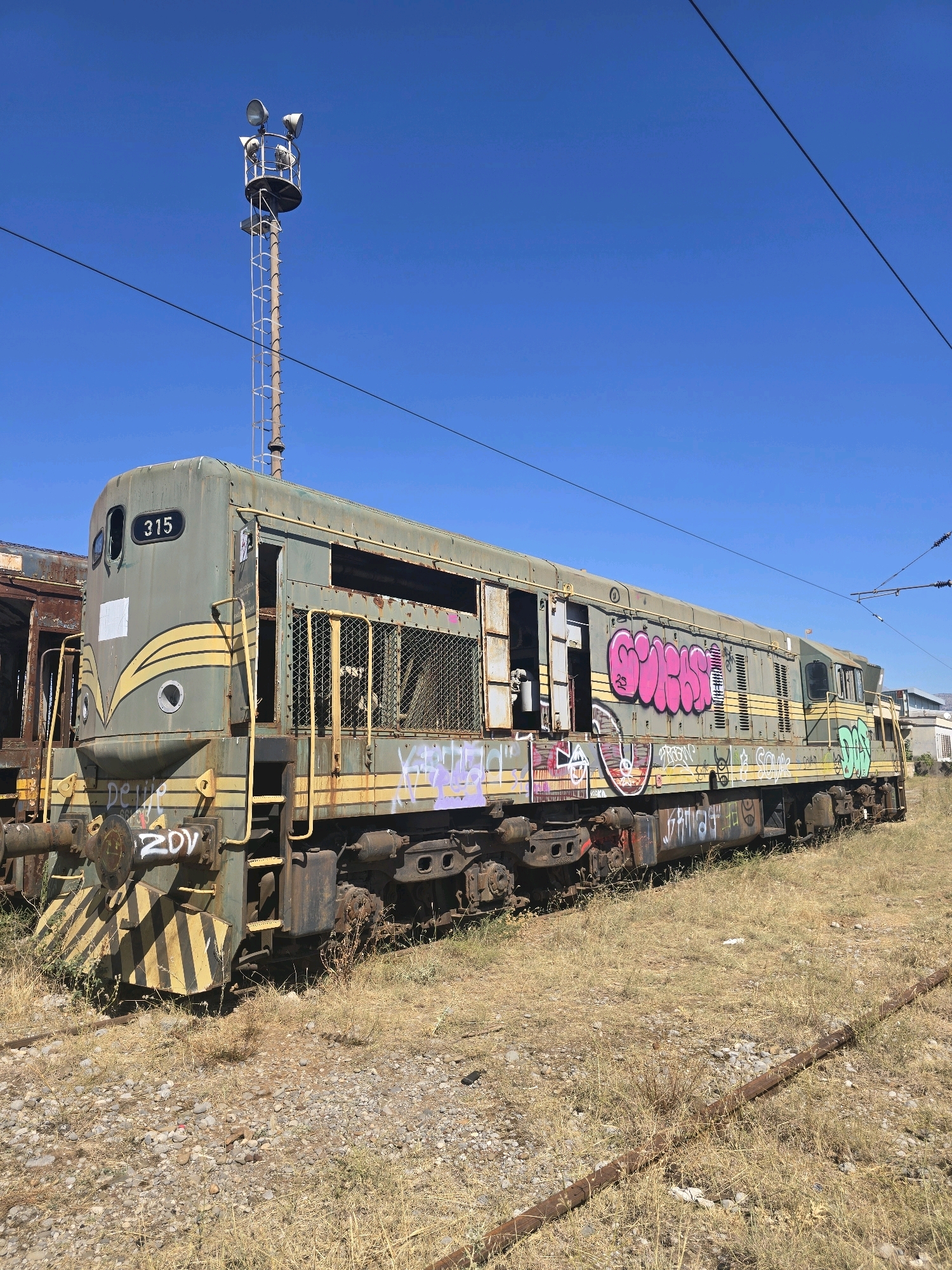 An abandoned, weathered, green diesel locomotive, numbered "315," is parked on disused train tracks in Podgorica, Montenegro. The locomotive is heavily tagged with graffiti in various colors, including pink, purple, and green. The exterior of the train is faded and rusted, with some windows broken or missing, and metal grates covering other openings. The locomotive is surrounded by dry, overgrown grass and gravel. In the background, a tall, metal pole with multiple spotlights stands against a clear blue sky. Overhead power lines are also visible. The overall scene suggests a derelict industrial area or a train yard that has fallen into disuse.