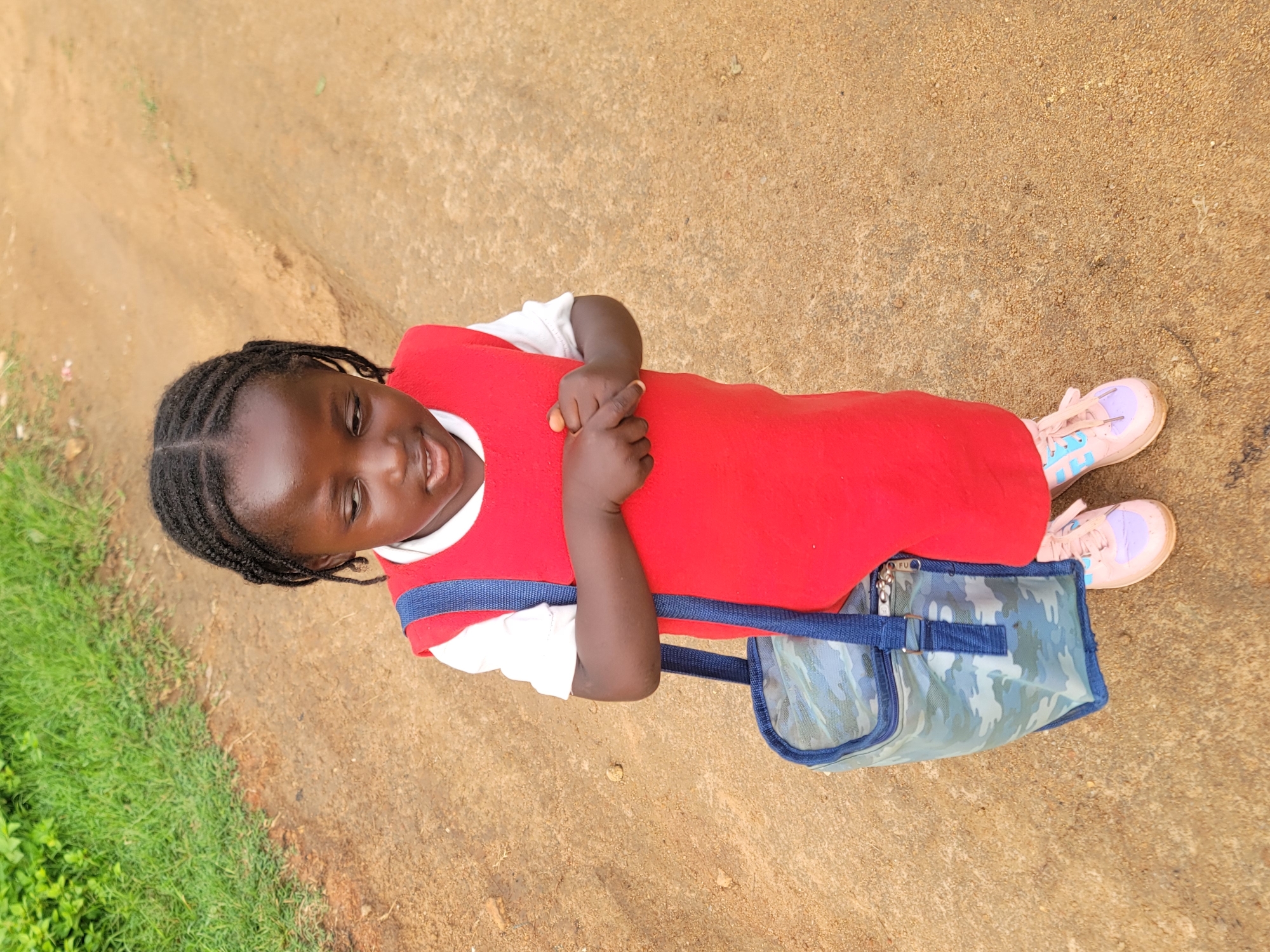 A young Black girl with braided hair stands on a dirt path, looking down. She is wearing a white shirt and a red sleeveless dress, with a camouflage bag slung over her left shoulder. Her hands are clasped in front of her. She is also wearing light pink and purple sneakers. To her left, a patch of green grass is visible. The background is a sandy, dirt path. This scene appears to be in the city of Barakin Akawo, Nigeria.