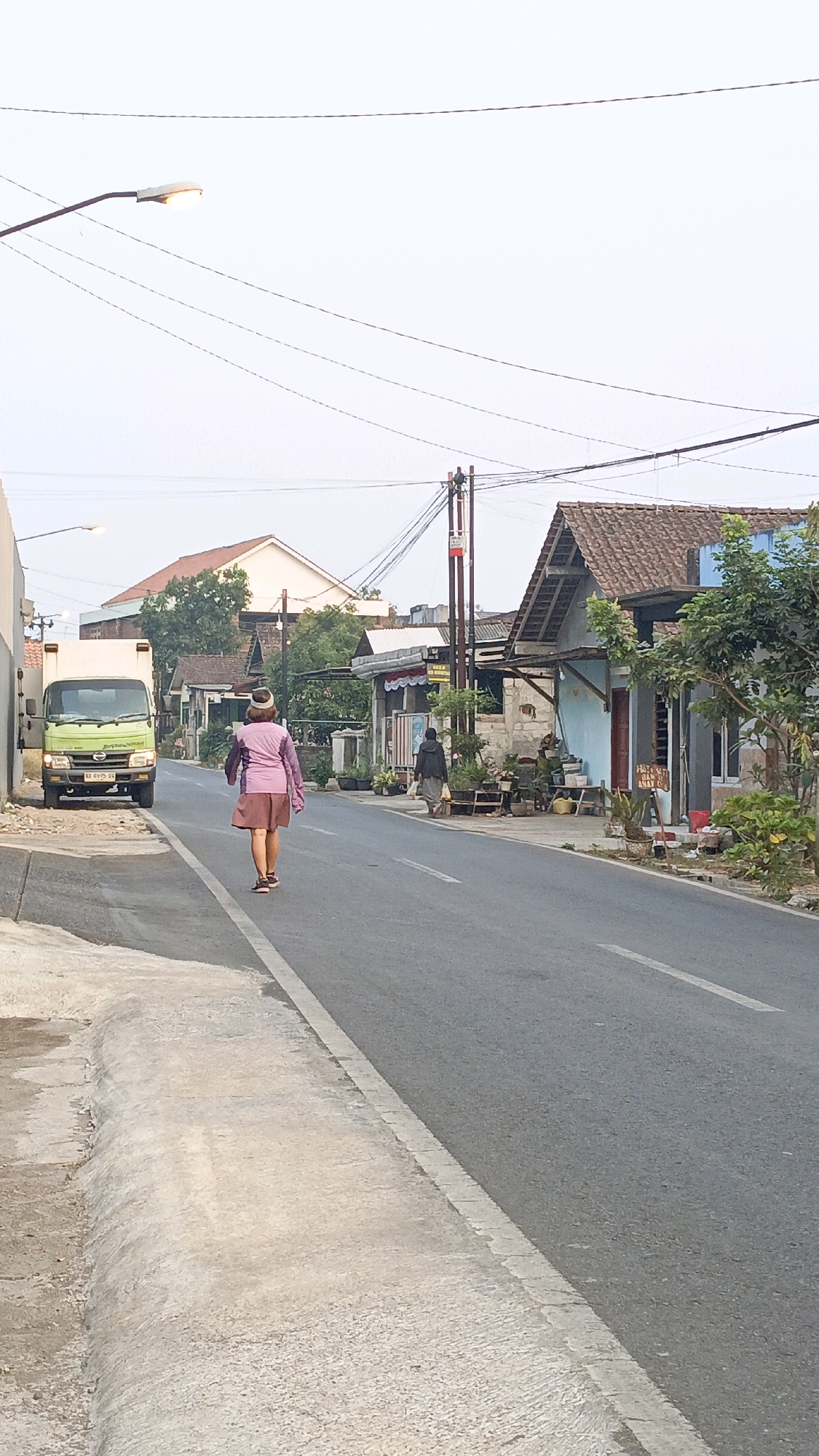 In Salatiga, Indonesia, a person walks away from the camera on the right side of an asphalt road. They are wearing a light pink jacket, a brown skirt, and a brown and white hat. In front of them, on the left side of the road, a light green truck is parked. The truck has a license plate that reads "AB 9016 GD". On the right side of the road, there are several buildings with tiled roofs, some with awnings and open fronts. Plants and potted flowers are visible in front of some of the buildings. The sky is overcast and light. Utility poles with wires run along the road. A street lamp is visible in the upper left corner of the image. The road has a white dashed line dividing the lanes. The sidewalk on the left is made of concrete and is slightly raised.