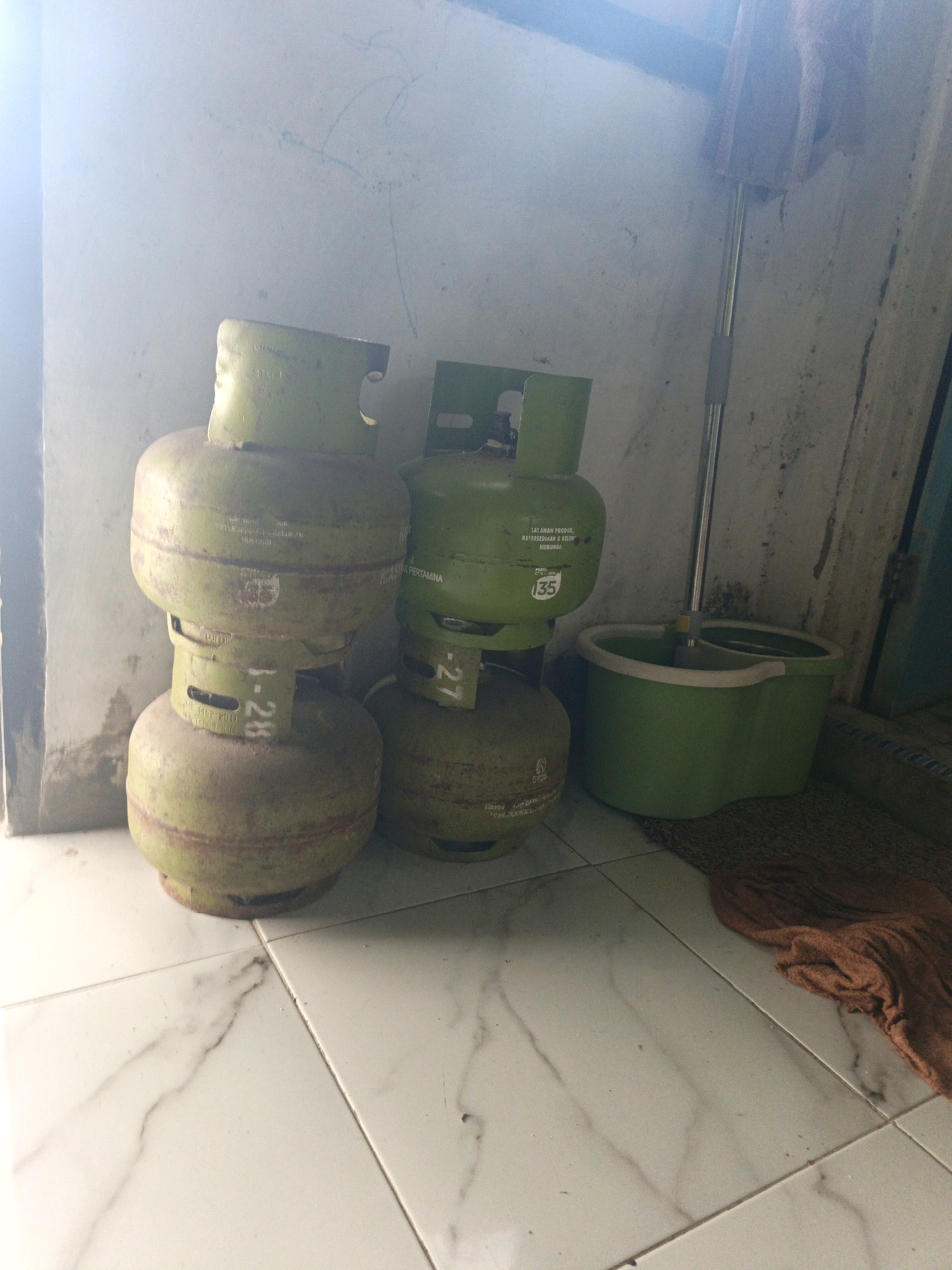 In Kamulyan, Indonesia, inside a room with white tiled flooring and a textured, off-white wall, three gas cylinders are stacked. The two bottom cylinders are a faded green, showing signs of wear and tear with some rust. The top cylinder on the left is a brighter green. The cylinder on the right, also green, is slightly behind the others. To the right of the cylinders, a green mop bucket sits on a brown mat, with a mop handle extending upwards and out of frame. A piece of orange fabric is draped over the right side of the mat. The lighting suggests either natural light from a window or a bright overhead source, casting soft shadows and creating a hazy effect in the upper left portion of the image. The wall has some faint blue marks, possibly child's drawings or graffiti. The overall scene depicts a storage area or corner of a room.