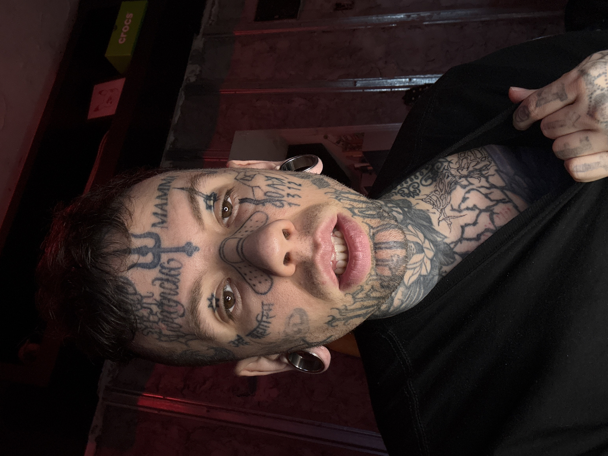 A male individual, appearing to be in his late twenties or early thirties, is depicted in a waist-up, frontal portrait in an indoor setting in Buenos Aires, Argentina. He has short, dark hair styled upwards. His face, neck, upper chest, and visible hands are extensively covered with black ink tattoos. Prominent facial tattoos include "Yoranda" and a trident symbol on the forehead, "MAMMA" above the right eyebrow, and a design resembling crossed adhesive bandages outlined on the nose. Text in a script, possibly Devanagari, is visible on the left cheek, while "80AX" and "1312" are tattooed on the right cheek. Both earlobes feature large, circular metal gauges. His brown eyes are fixed on the camera, and his lips are slightly parted, revealing his upper teeth. He wears a plain black short-sleeved garment, which he pulls down with his tattooed right hand to expose densely patterned abstract tattoos on his upper chest. The background is dimly lit with a reddish hue. A dark shelf in the upper right displays a green box labeled "crocs" and a smaller red item. The wall behind the subject appears to have a textured or paneled surface.