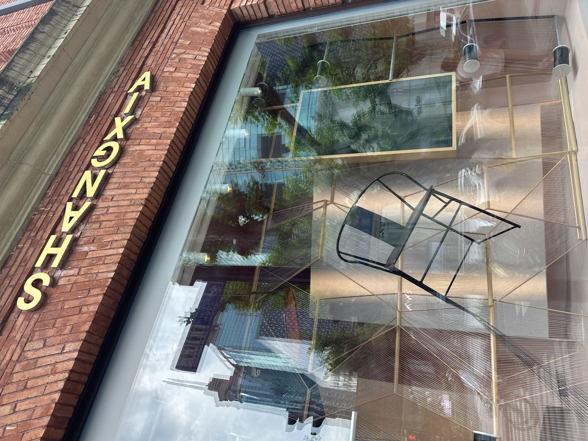 The image shows a storefront with the name "SHANGHAI" in gold lettering above a large glass window. The window reflects buildings and trees, and inside the store, there is abstract geometric decor and a black chair. The storefront is made of red brick.