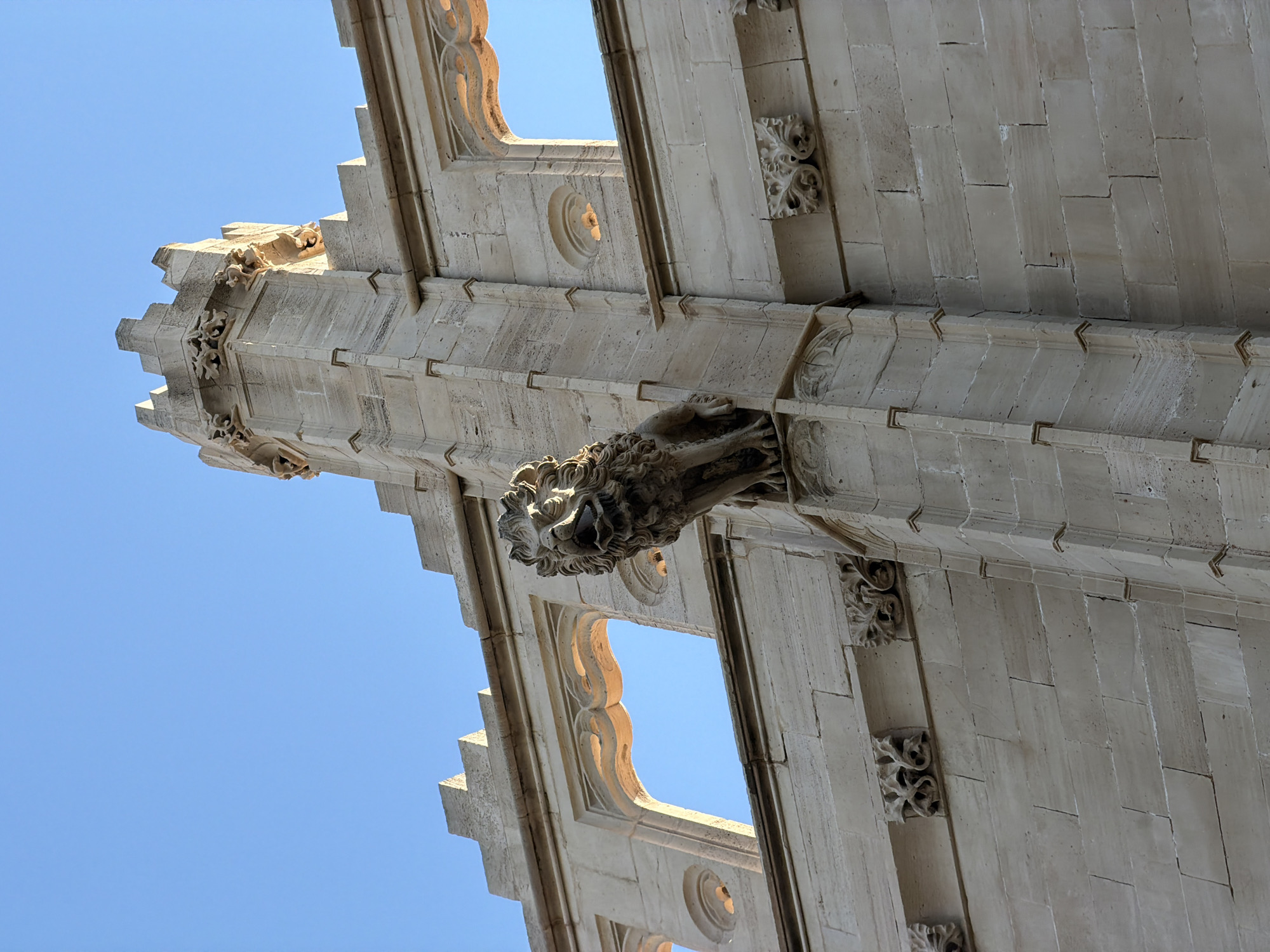 The image shows a close-up, low-angle view of a stone gargoyle shaped like a lion on the side of a building, with a clear blue sky in the background. The gargoyle is positioned on a corner of the building, with its mouth open and mane detailed. The building itself is constructed of light-colored stone blocks, with architectural details including arched windows and decorative carvings. The style of the architecture appears Gothic or Romanesque. Based on the distinctive architectural style and the presence of gargoyles of this nature, the building is identified as the Cathedral of Santa Maria of Palma, also known as La Seu, located in Palma de Mallorca, Spain. The gargoyle is a functional architectural element designed to divert rainwater away from the building's walls. There are no people or other living creatures visible in the image. The lighting suggests it is daytime, with sunlight illuminating the stone.