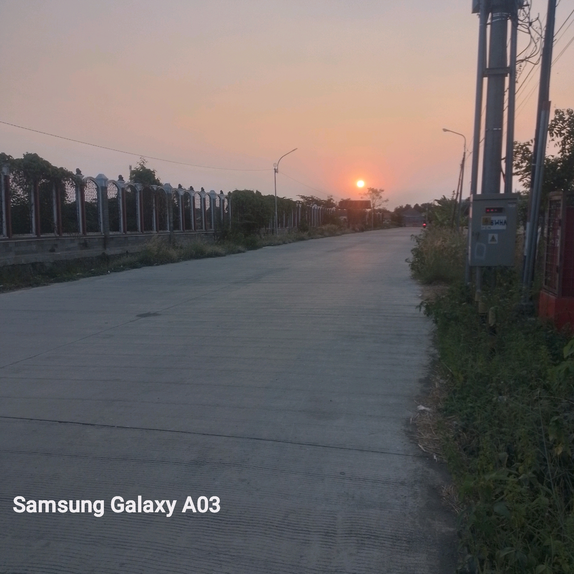 This is a landscape photograph of a road at sunset, likely in Indramayu, Indonesia, as indicated by the text overlay "Samsung Galaxy A03". The setting sun casts a warm, orange glow across the sky, with the sun itself visible as a bright orb near the horizon. A paved road stretches into the distance, bordered on the left by a decorative fence with arched panels and lush green foliage. On the right side of the road, there is an electrical pole with various wires and a gray electrical box mounted on it. The foreground shows the texture of the asphalt road. The overall atmosphere is serene and tranquil, characteristic of a rural or suburban environment at the end of the day.