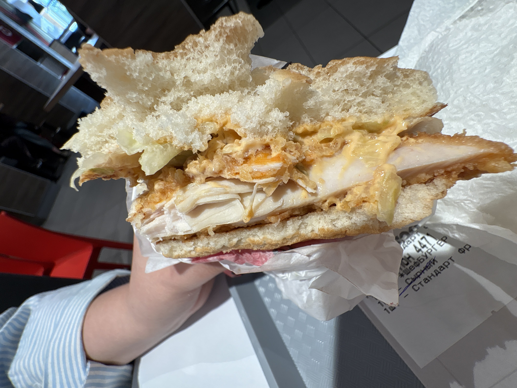 A partially eaten chicken sandwich is held in a person's hand. The sandwich consists of a white, textured bun with chicken breast slices, shredded lettuce, and a creamy sauce. The sandwich is wrapped in white paper that has Russian writing on it. The paper reads in Cyrillic script: "CH 447 Шевбургер Сырный Стандарт ФР". This translates to "CH 447 Shefburger Cheese Standard FR". The person holding the sandwich is wearing a blue and white striped shirt. A gray tray is visible underneath the paper. A red chair is partially visible in the blurred background. Based on the product packaging, the location is likely Izhevsk, Russia.