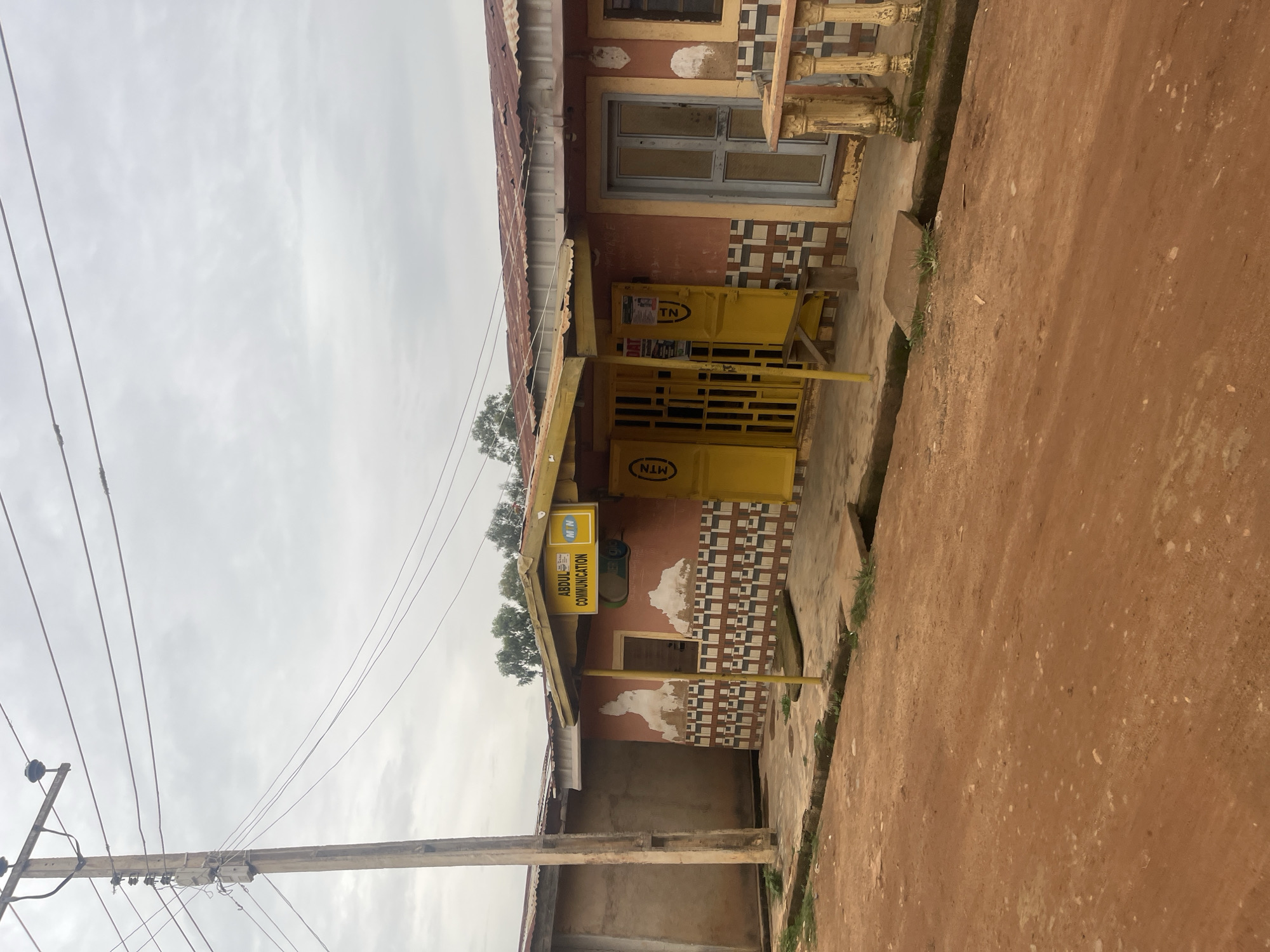 A medium shot captures a small shopfront in what appears to be Bukuru, Nigeria, on an overcast day. The shop, painted a reddish-orange, features a yellow awning supported by a yellow pole. A yellow sign above the entrance reads "ABDUL COMMUNICATION." The shop's facade is decorated with a repetitive geometric pattern in brown and beige. Two yellow, metal-barred doors are visible. To the right of the shop, a light blue door is set into a reddish-orange wall, flanked by a decorative yellow railing with vertical supports. A concrete sidewalk runs in front of the shop, with a dirt road in the foreground. A wooden utility pole with electrical wires stands to the left of the shop. The background shows the corrugated metal roof of the shop and adjacent buildings, along with some greenery. No people are present in the scene.