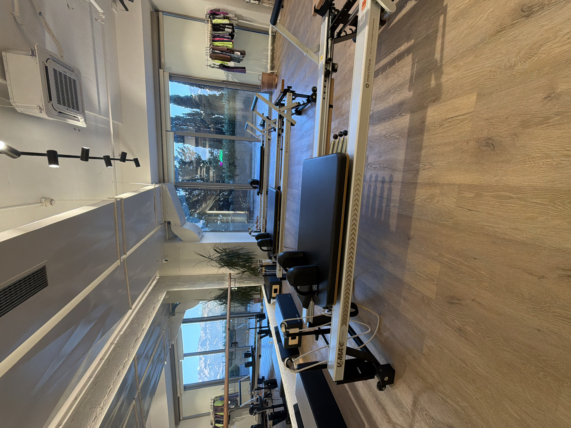 The image depicts the interior of a Pilates studio located in Boreti, Montenegro. The room features several Pilates reformers, which are exercise machines consisting of a carriage that slides back and forth on a track, supported by a frame. These reformers are arranged in parallel rows on a polished wooden floor. Large windows at the back of the room offer a view of snow-capped mountains and trees, suggesting a scenic location.
In the foreground, a white and grey Pilates reformer is prominently displayed. It has a black padded carriage with headrest and shoulder rests. The side of the reformer is marked with the brand name "V-MAX REFORMER" and "merrithew leaders in mindful movement." Additional reformers are visible further back in the room, some appearing to be folded or in a vertical storage position.
A rack of athletic clothing, including leggings and tops in various colors, hangs on a rack against the wall to the right of the windows. A wicker basket is placed on the floor near the clothing rack. The ceiling features exposed beams, lighting fixtures, and an air conditioning unit. The overall ambiance suggests a clean, modern, and well-equipped fitness space. No people are visible in the image.