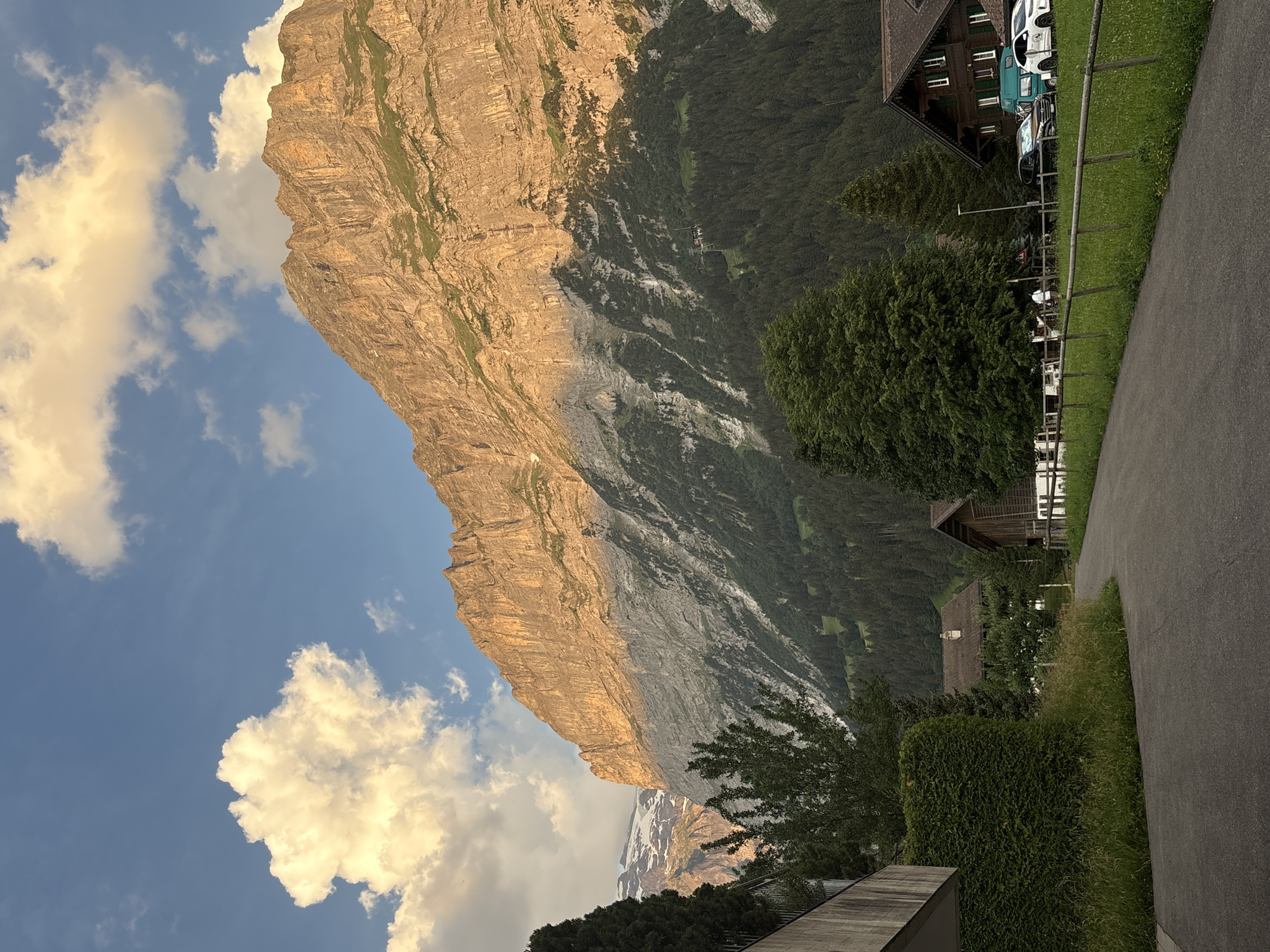 The image shows a view of a mountain in Grindelwald, Switzerland, bathed in the warm glow of the setting sun. In the foreground, a paved road curves to the right, bordered by green grass and a wooden fence. To the left of the road, a modern concrete building with a balcony is visible, partially covered by a manicured hedge. Further back, traditional Swiss chalets with dark wooden siding and gabled roofs are nestled amongst trees. A small dark-colored car and a white SUV are parked in front of one of the chalets. The mid-ground is dominated by a dense forest of evergreen trees that ascend the lower slopes of the mountain. The mountain itself is a massive, imposing structure, with its upper reaches illuminated by the golden light of the sun, creating dramatic shadows and highlighting its rugged texture. Patches of green grass cling to the lower slopes, transitioning to bare rock and scree higher up. The sky above is a vibrant blue, dotted with fluffy white and yellow clouds, some of which are also catching the sunlight. In the distance, another snow-capped mountain peak can be seen to the left. The overall scene evokes a sense of serene natural beauty and the charm of a Swiss alpine village.