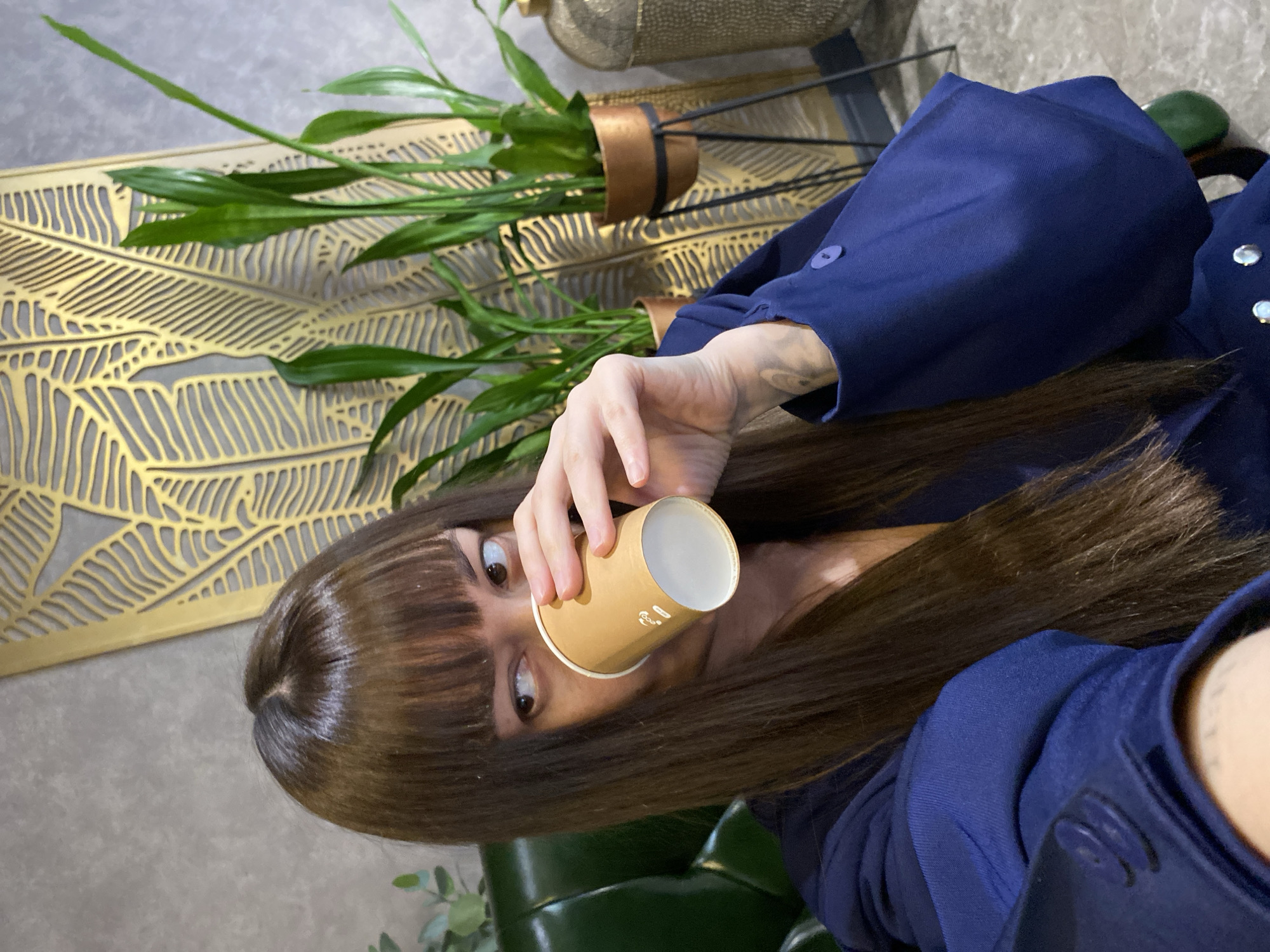 A woman is photographed indoors, angled to the side, drinking from a brown paper cup. She has brown hair with bangs and is wearing a blue garment with buttons. Behind her are plants in copper-colored pots and a decorative gold-colored wall panel with a leaf pattern. The location and city, Votkinsk, Russia, are not verifiable from the image.