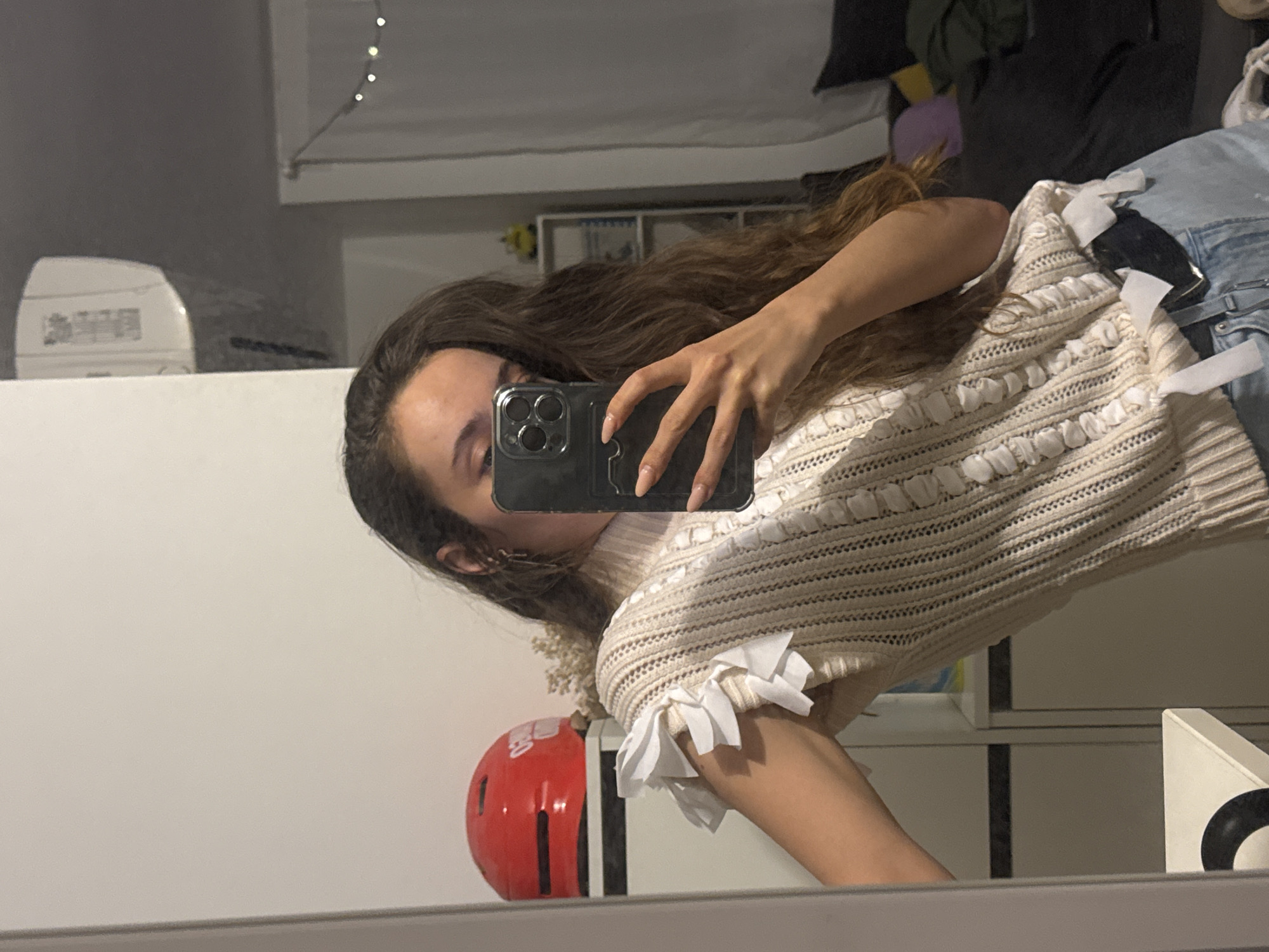 A person stands in front of a mirror, holding a smartphone in their right hand to take a selfie. The phone obscures most of the person's face except for the eyes, eyebrows, and part of the nose. The person has long, wavy brown hair. They wear a beige-colored, horizontally-striped sweater with ruffled white fabric accents. The sweater sits above a pair of light-wash denim jeans.
Behind the person and reflected in the mirror is a room. A red helmet rests on a surface near the bottom of the frame. To the left of the person in the mirror is a white object on top of a white counter or shelf. Above that, there is a string of lights. Other objects and clothing are present in the background.
The scene is indoors, with no discernible indication of a specific location within Ramat Gan, Israel. No specific actions or interactions are evident beyond the person taking the selfie.